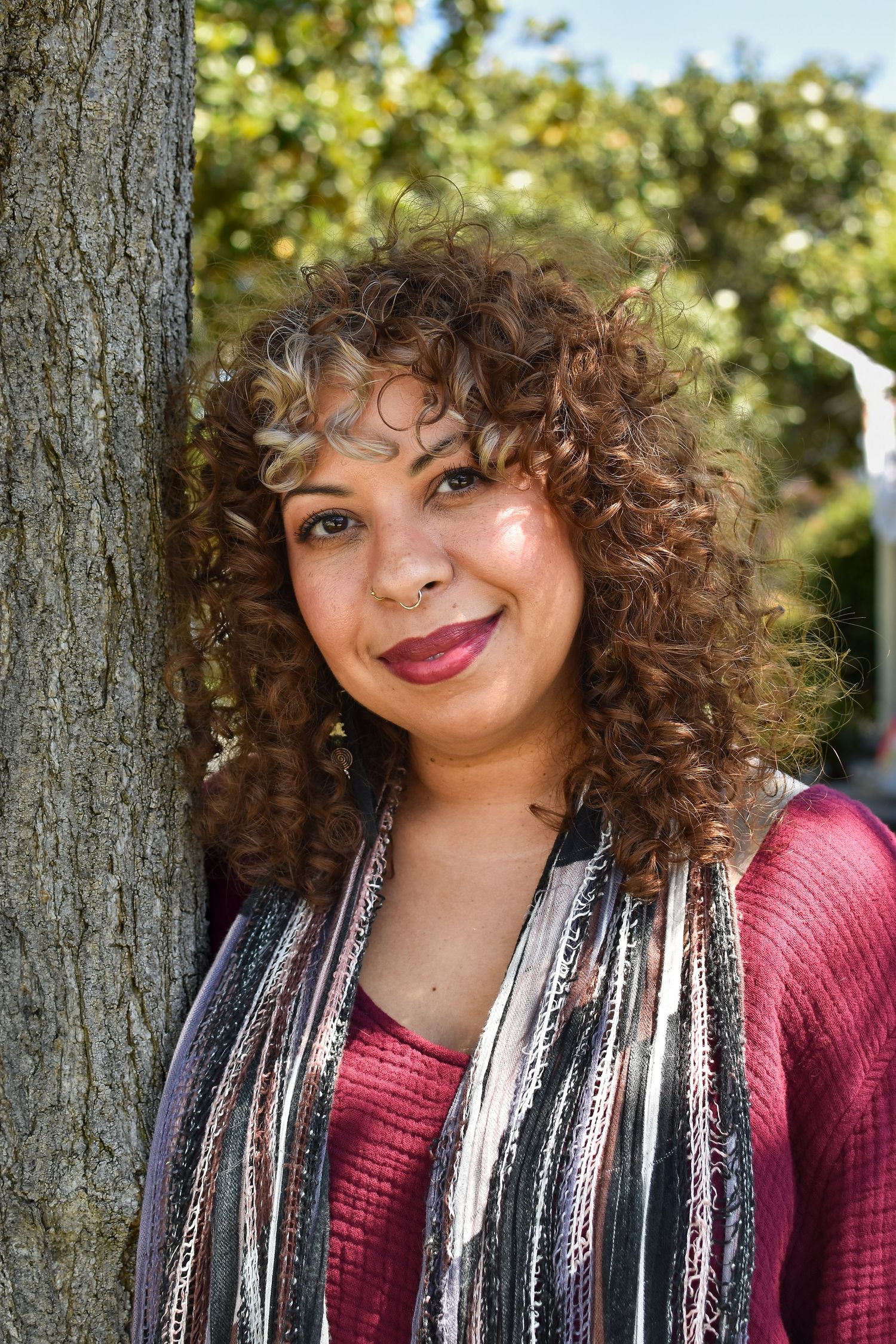 Portrait of someone with natural curly hair wearing a red top and striped scarf leaning against a tree trunk outdoors.