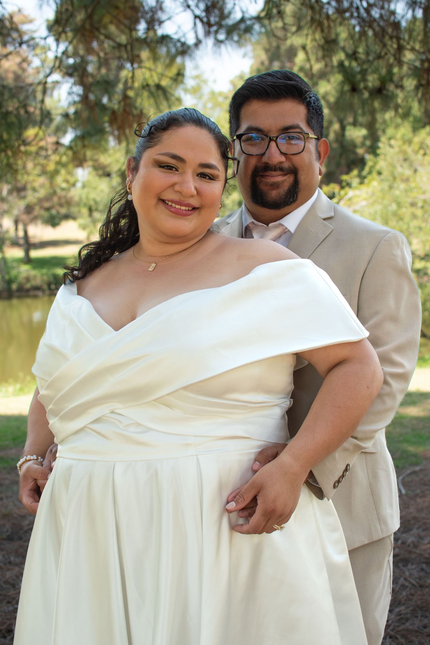 A happy couple in wedding attire pose together outdoors with trees in the background on their special day.