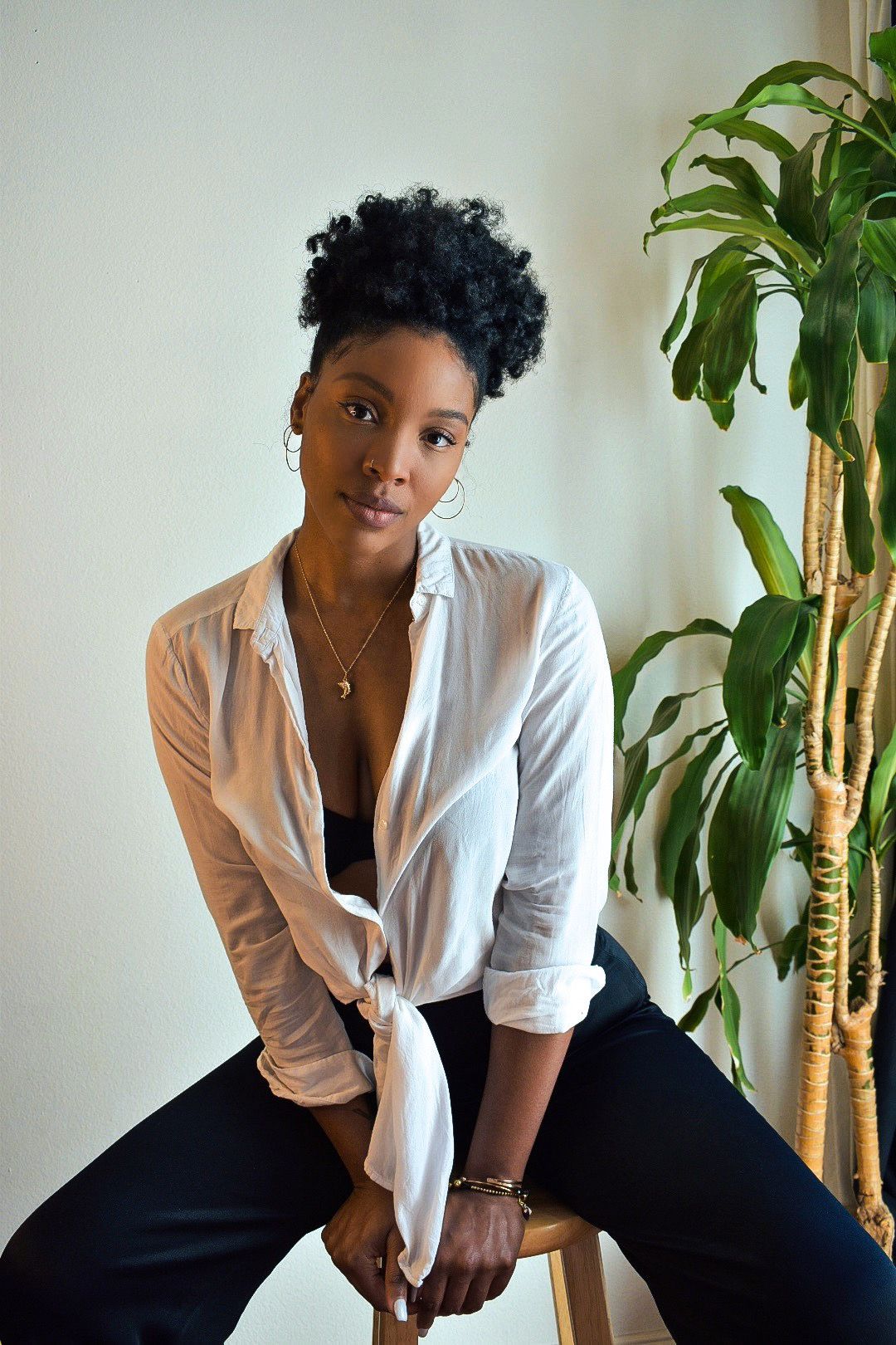 A person in a white button-down shirt and black pants poses casually near a large indoor plant against a light wall.