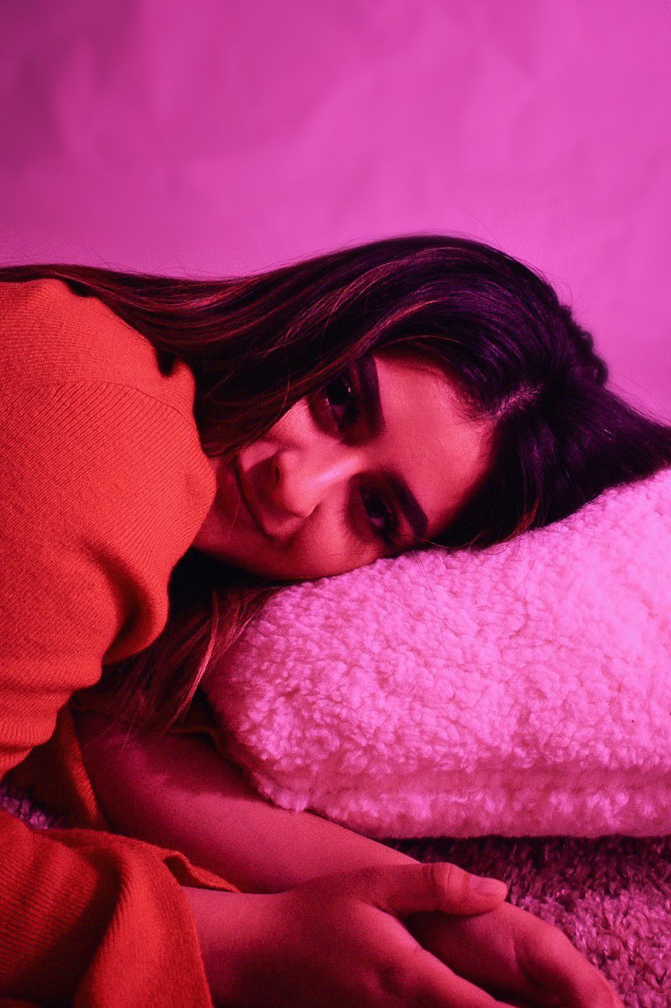 Dreamy portrait against pink backdrop with figure in red sweater reclining on soft surface.