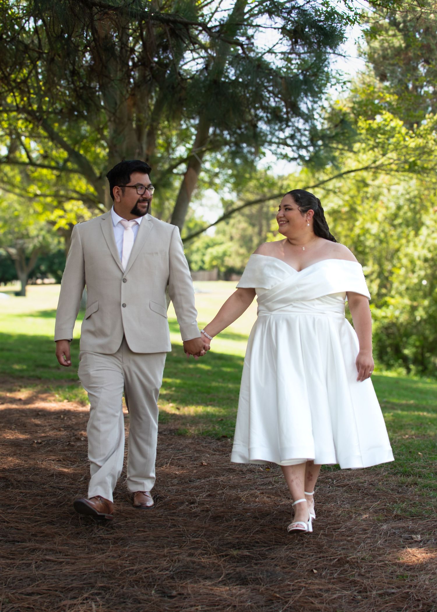 A couple in a beige suit and white dress holding hands while standing together on a wooded path in a park setting.