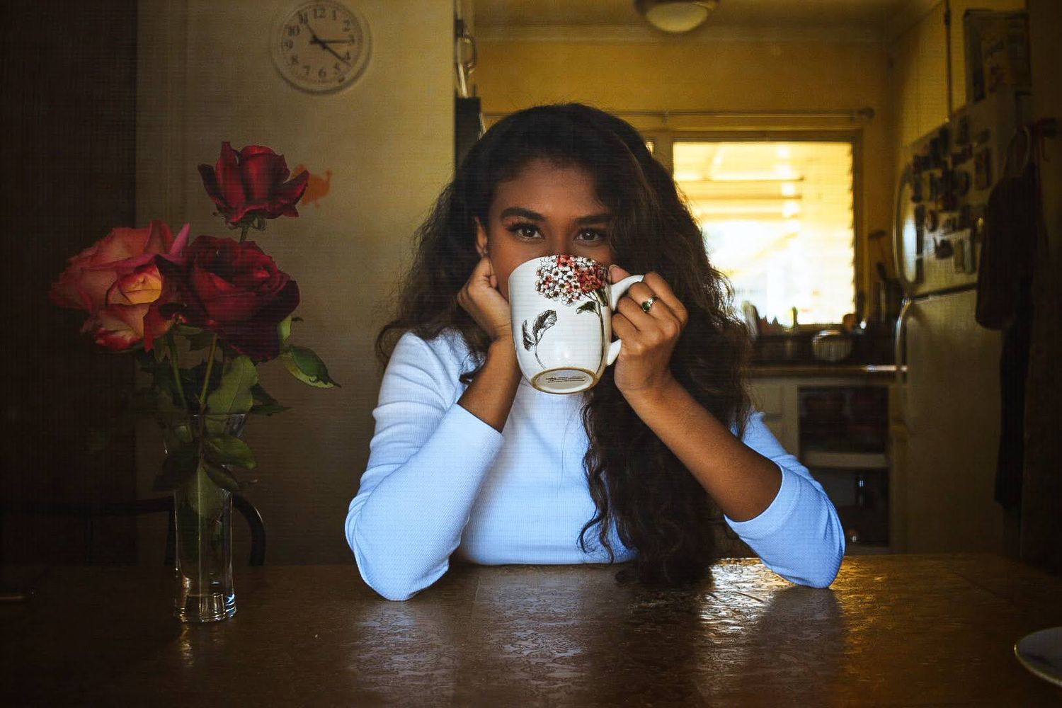 Person sipping from a decorative mug at a wooden table with red roses in the background.