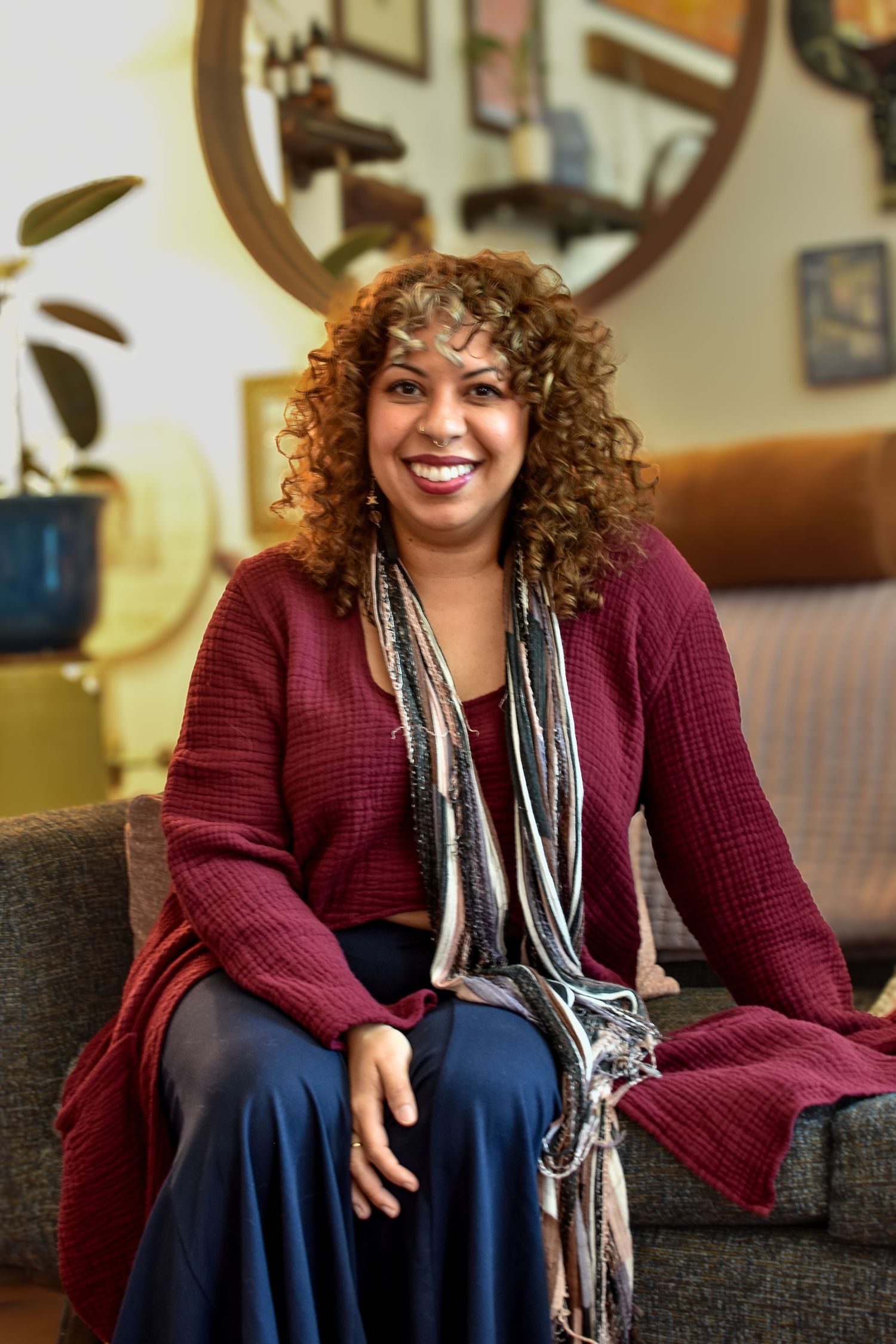 Someone in a burgundy cardigan and striped scarf sits casually on a dark gray couch with a warm smile.
