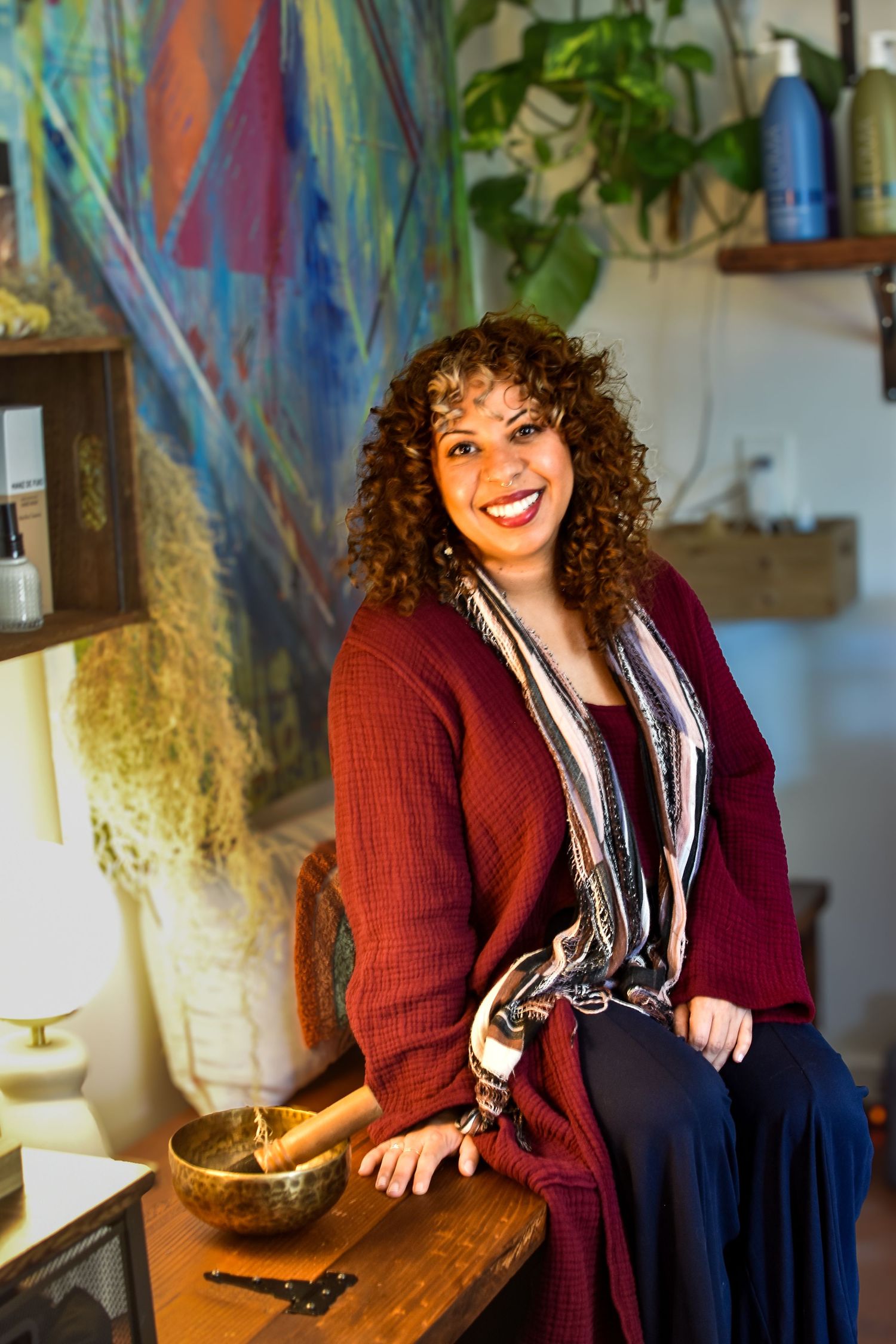 A person wearing a burgundy cardigan and striped scarf sits next to a singing bowl in a cozy room with colorful artwork.
