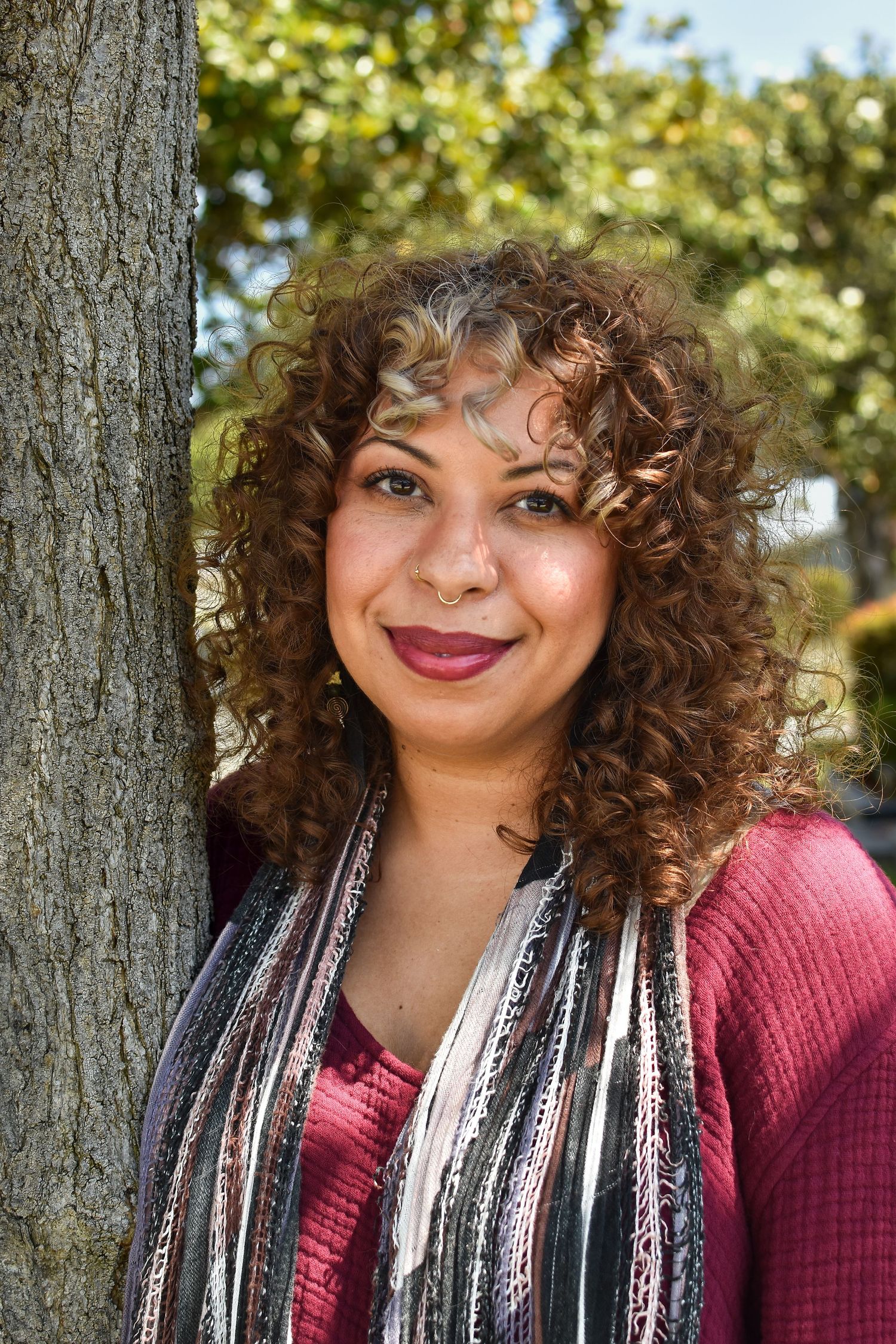 A person with natural curly hair leans against a tree trunk while wearing a burgundy sweater and striped scarf outdoors.