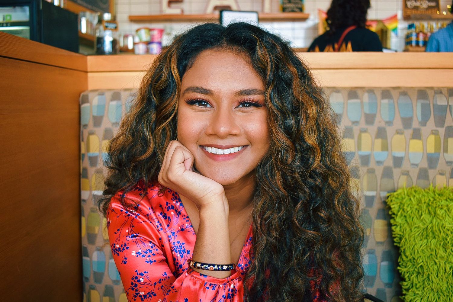 Portrait of someone with curly hair smiling and resting chin on hand in casual restaurant setting.