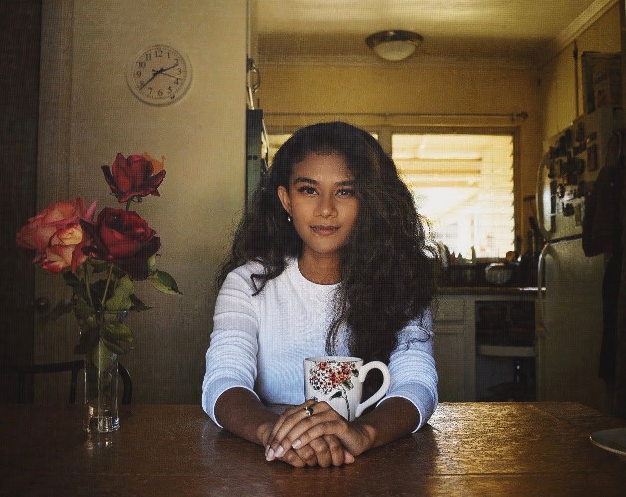 Person seated at wooden table with patterned mug and red roses in moody lighting.