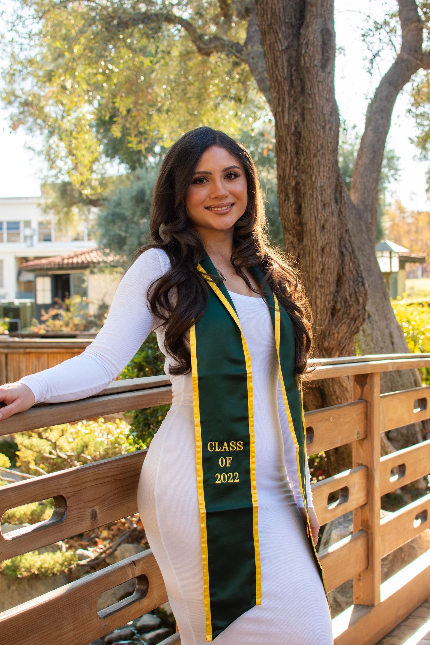 Graduate in white dress and green sash poses on wooden bridge during graduation photoshoot.