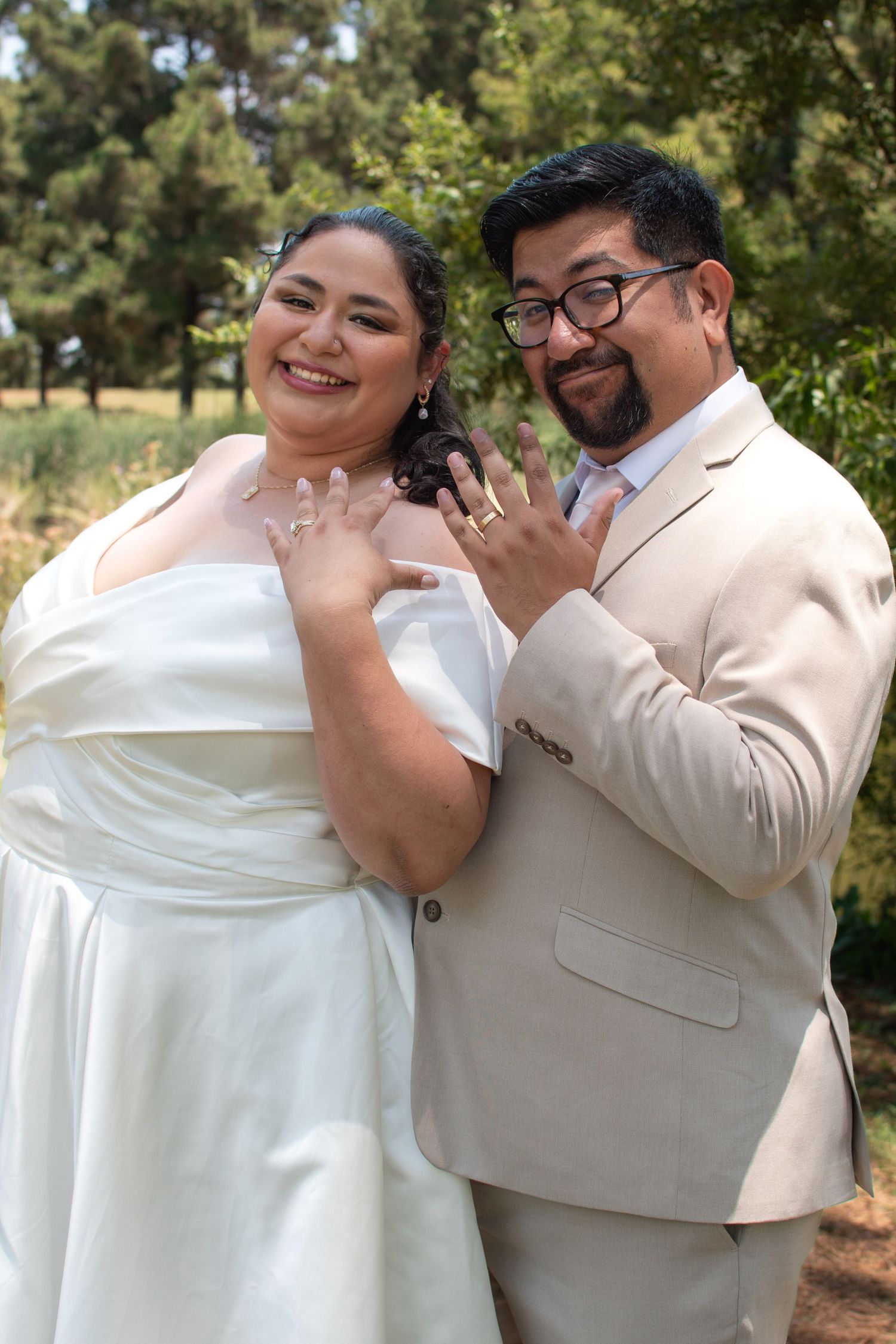 A couple shares a joyful moment together outdoors, wearing white and beige formal attire in a natural setting.