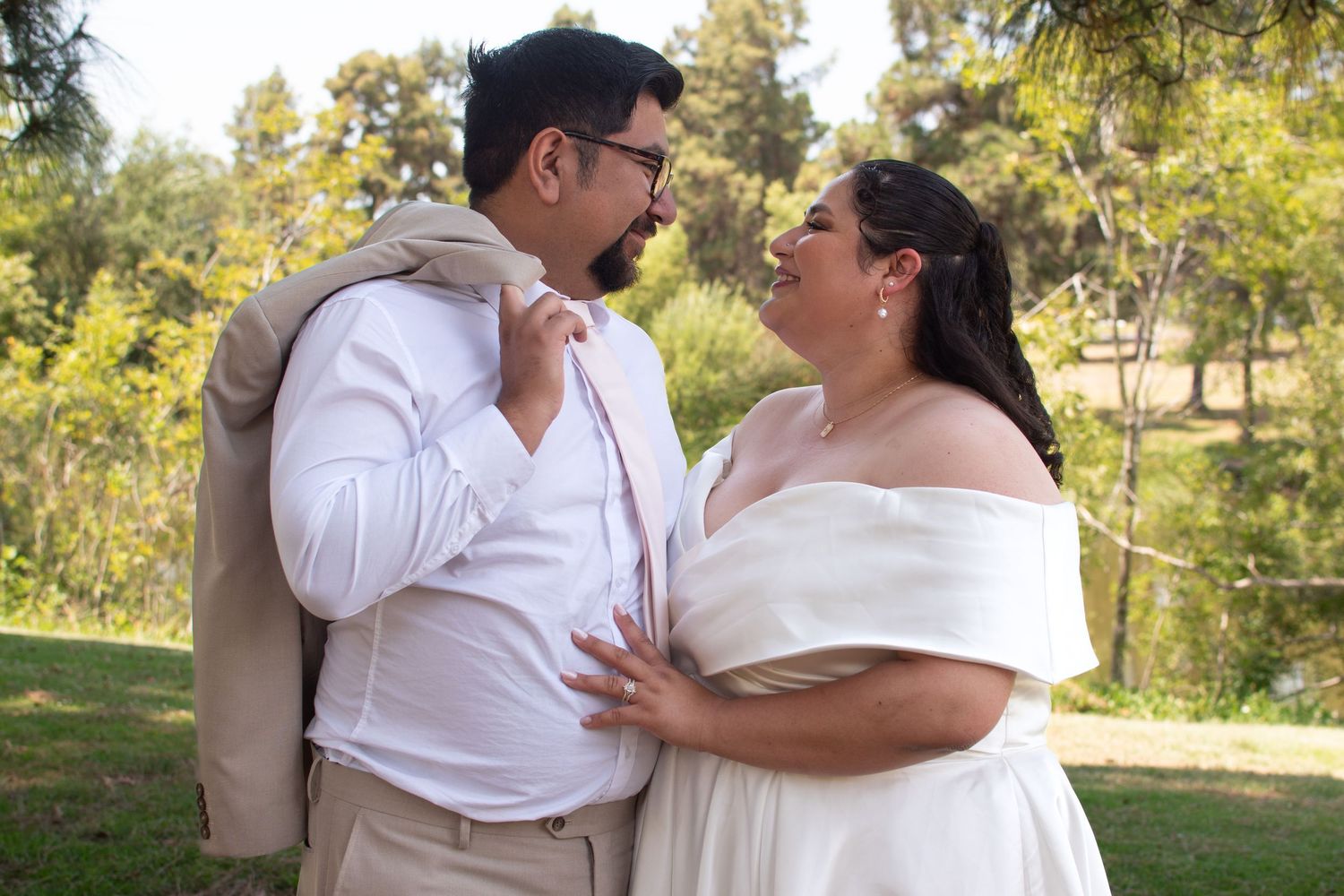 A romantic couple in white attire share an intimate moment together outdoors in a sunlit garden setting.