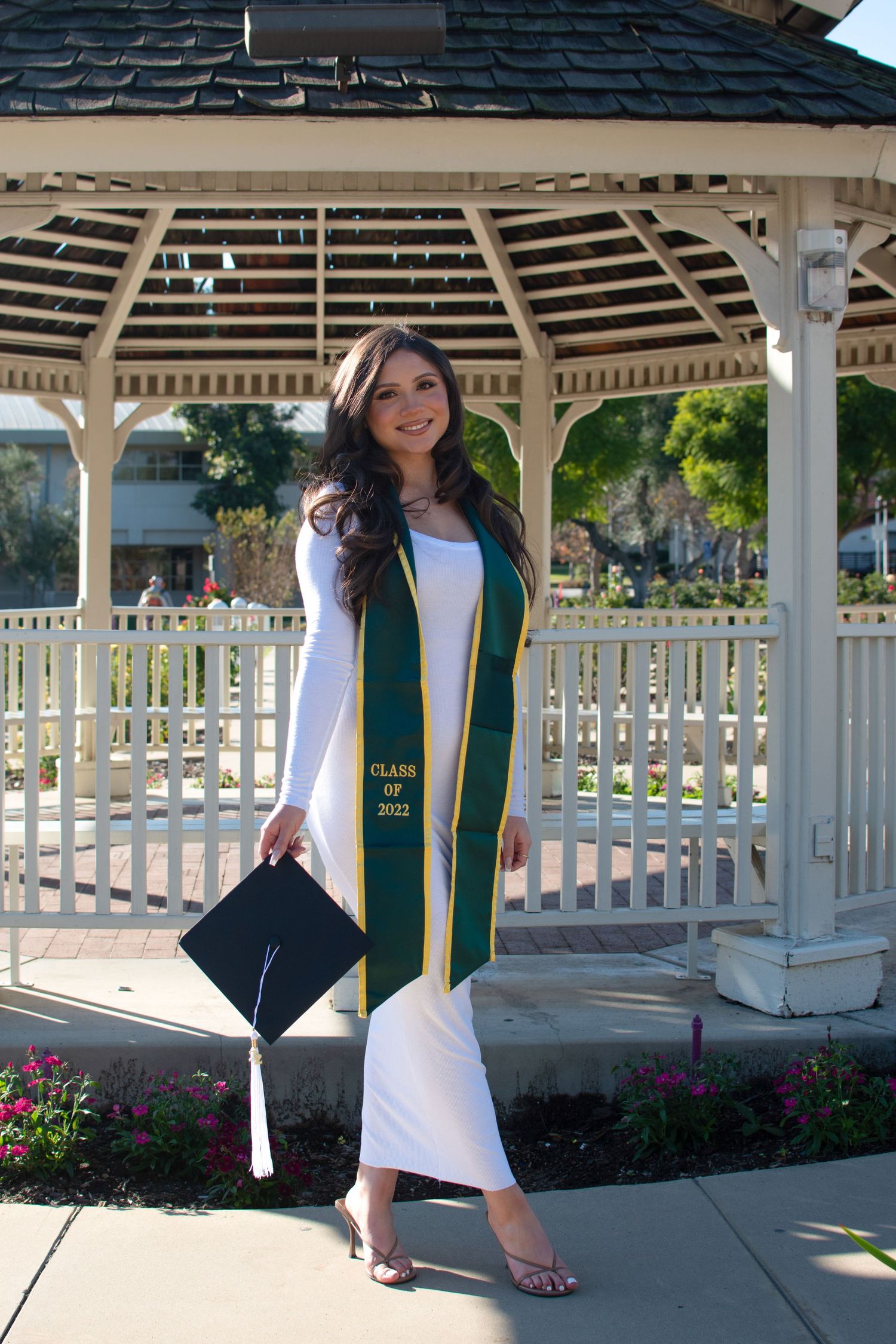 Graduate in white dress and green sash poses in outdoor gazebo for graduation photos.