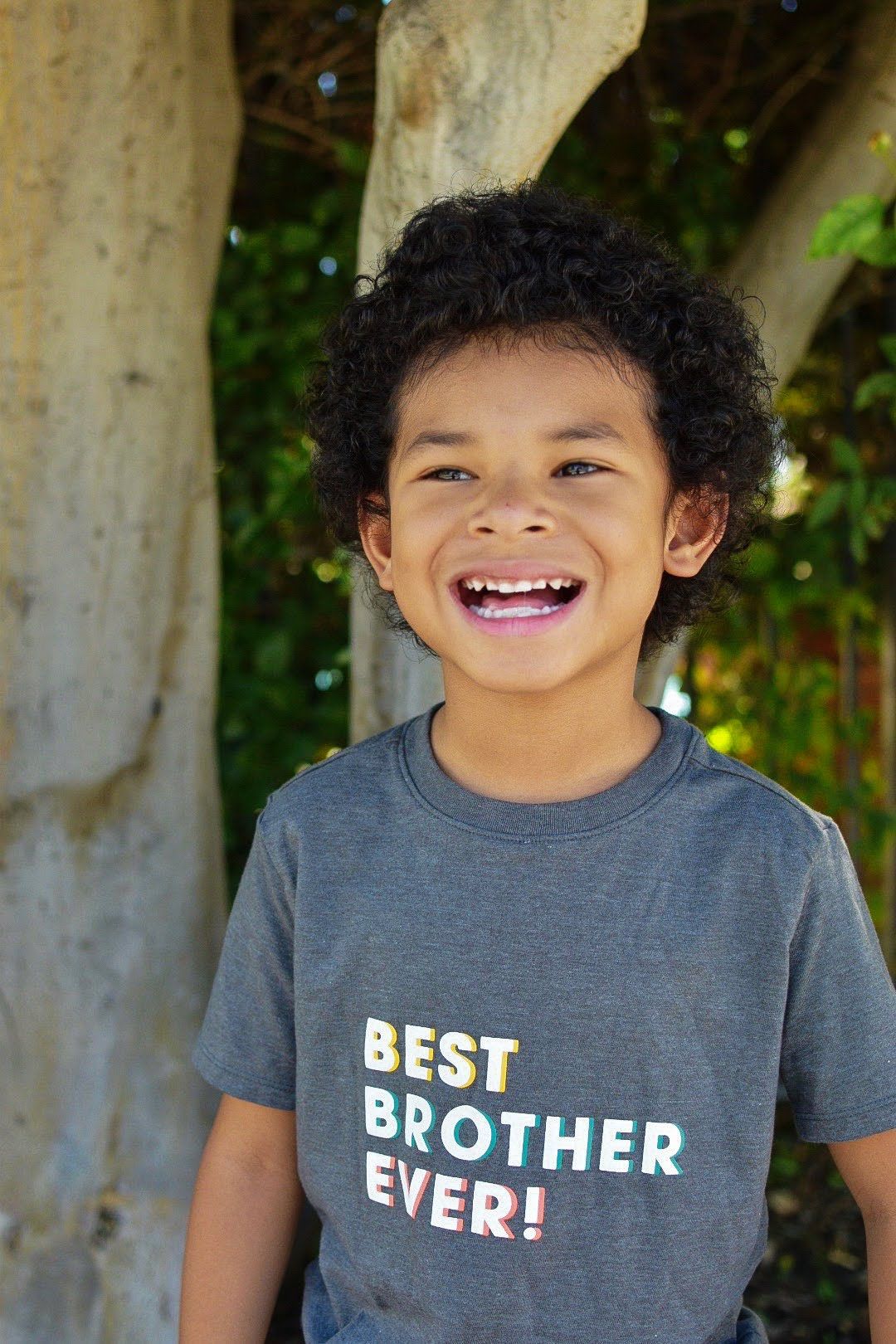 Child wearing grey t-shirt with 'Best Brother Ever' text smiles brightly outdoors.