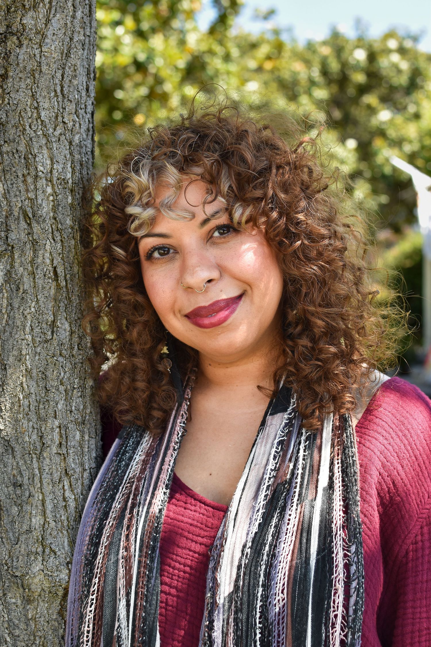 A casual outdoor portrait showing curly hair and a burgundy top against a natural tree background.