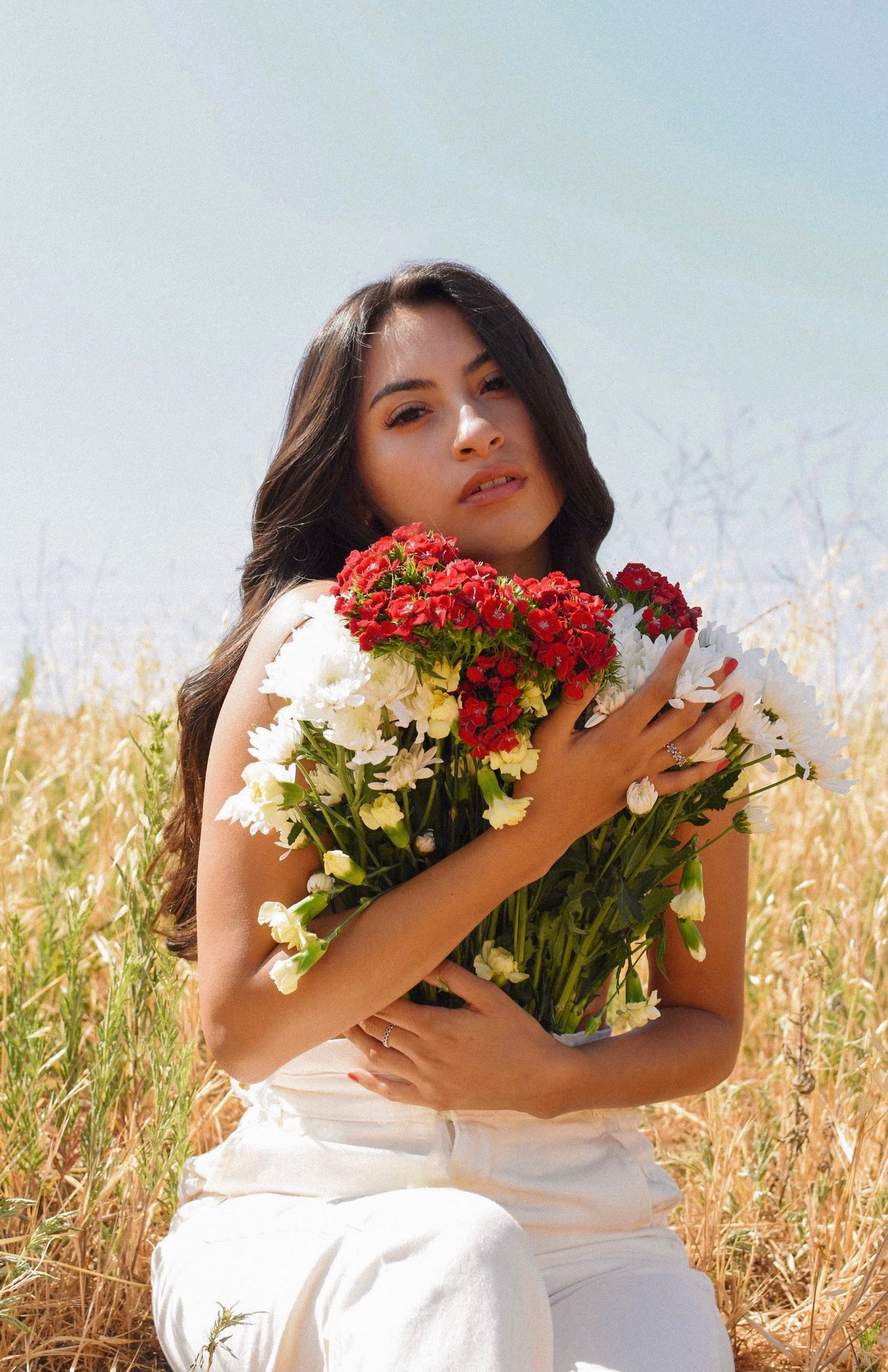 Woman embracing bouquet of red and white flowers against sunny wheat field background.