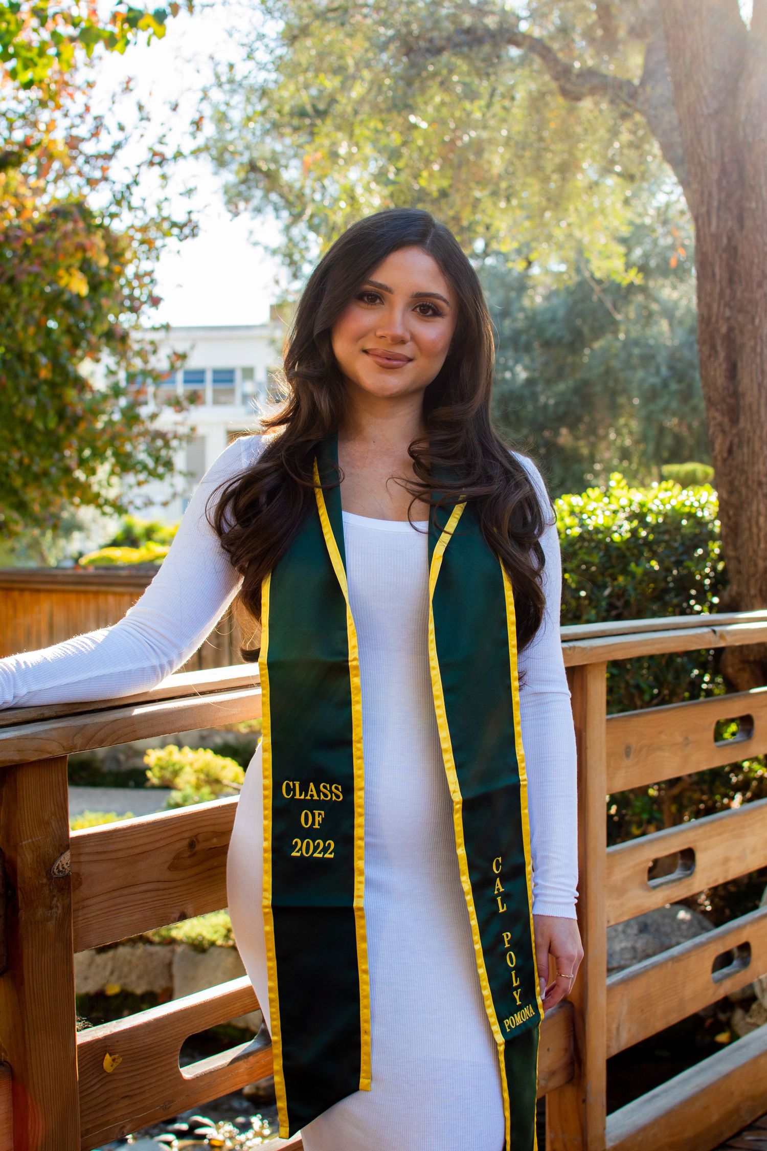 Graduate in white dress with Class of 2022 stole poses on wooden bridge in natural setting.