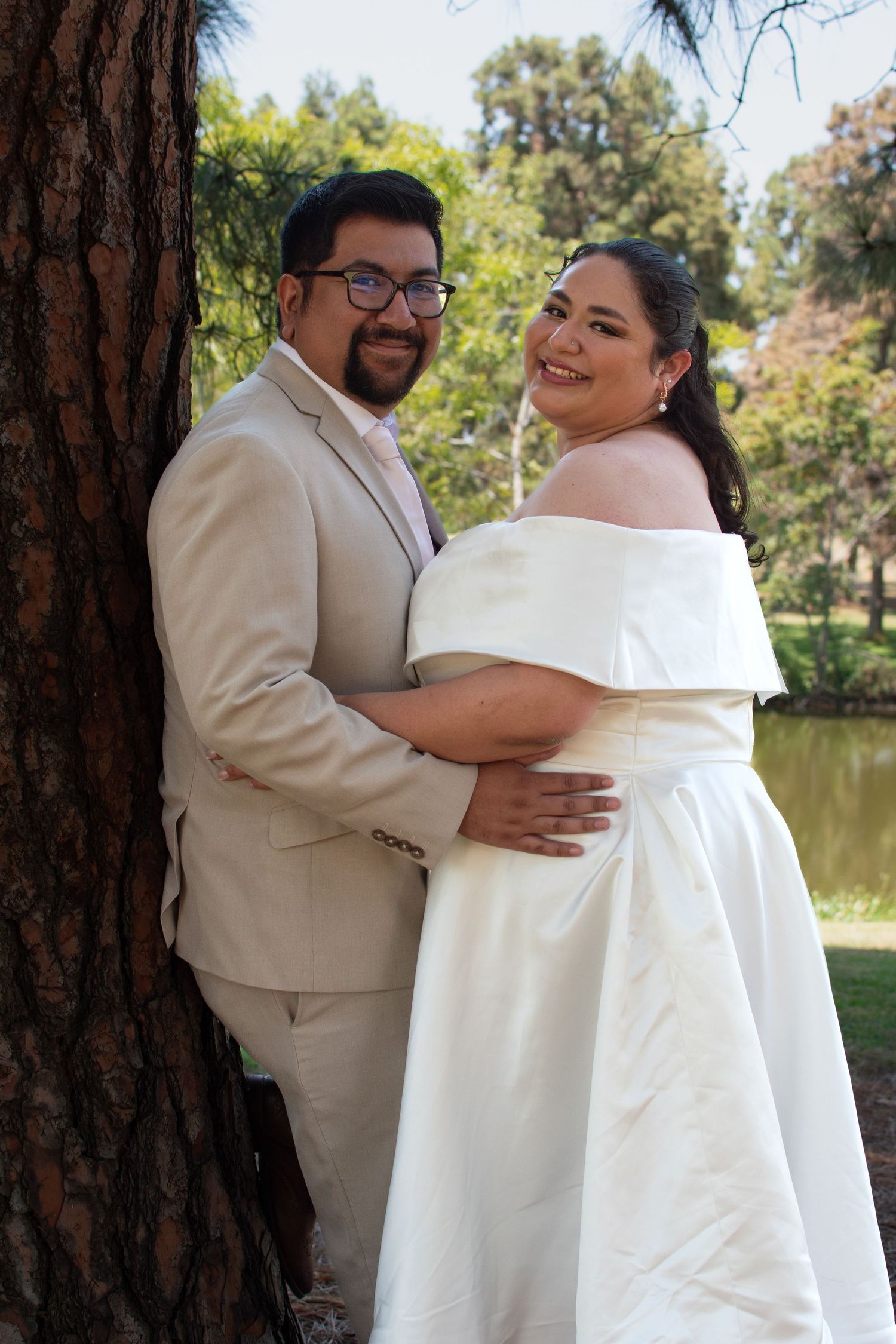 A couple poses together by a tree next to a lake, wearing matching beige and white formal wedding attire.