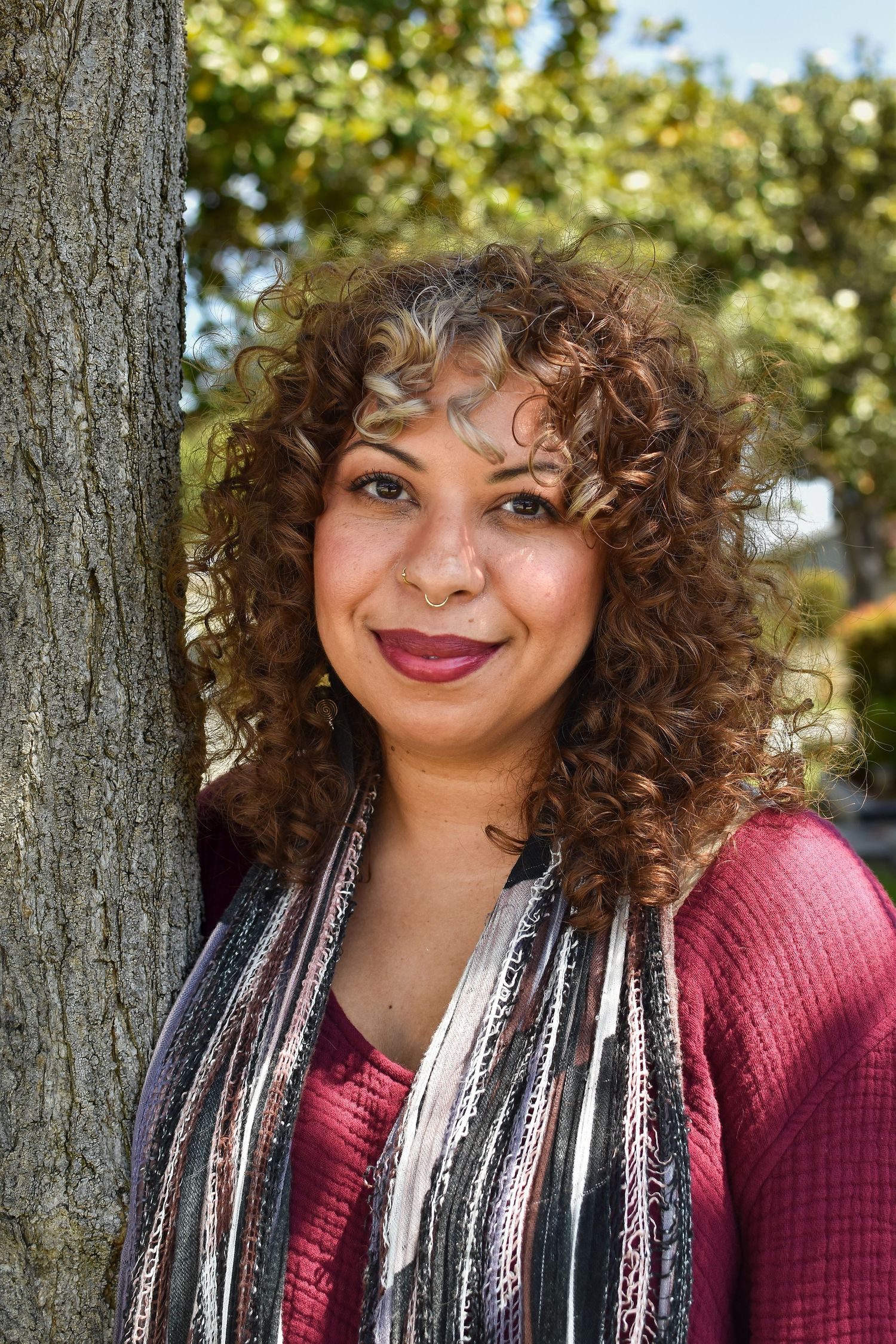 Portrait of someone with curly hair wearing a burgundy sweater and patterned scarf leaning against a tree trunk outdoors.