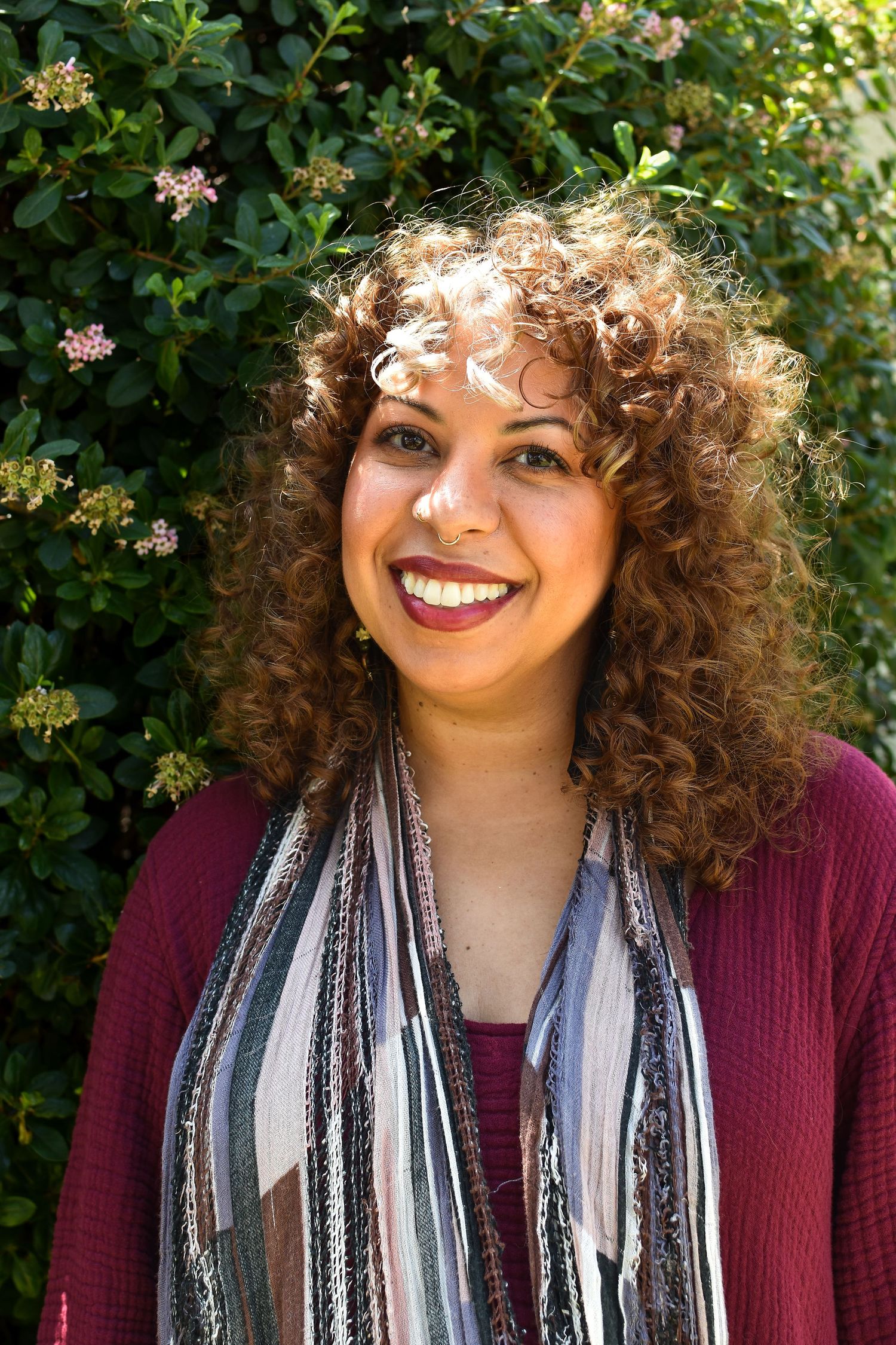 A portrait outdoors against greenery wearing a burgundy cardigan and patterned scarf with natural curly hair.