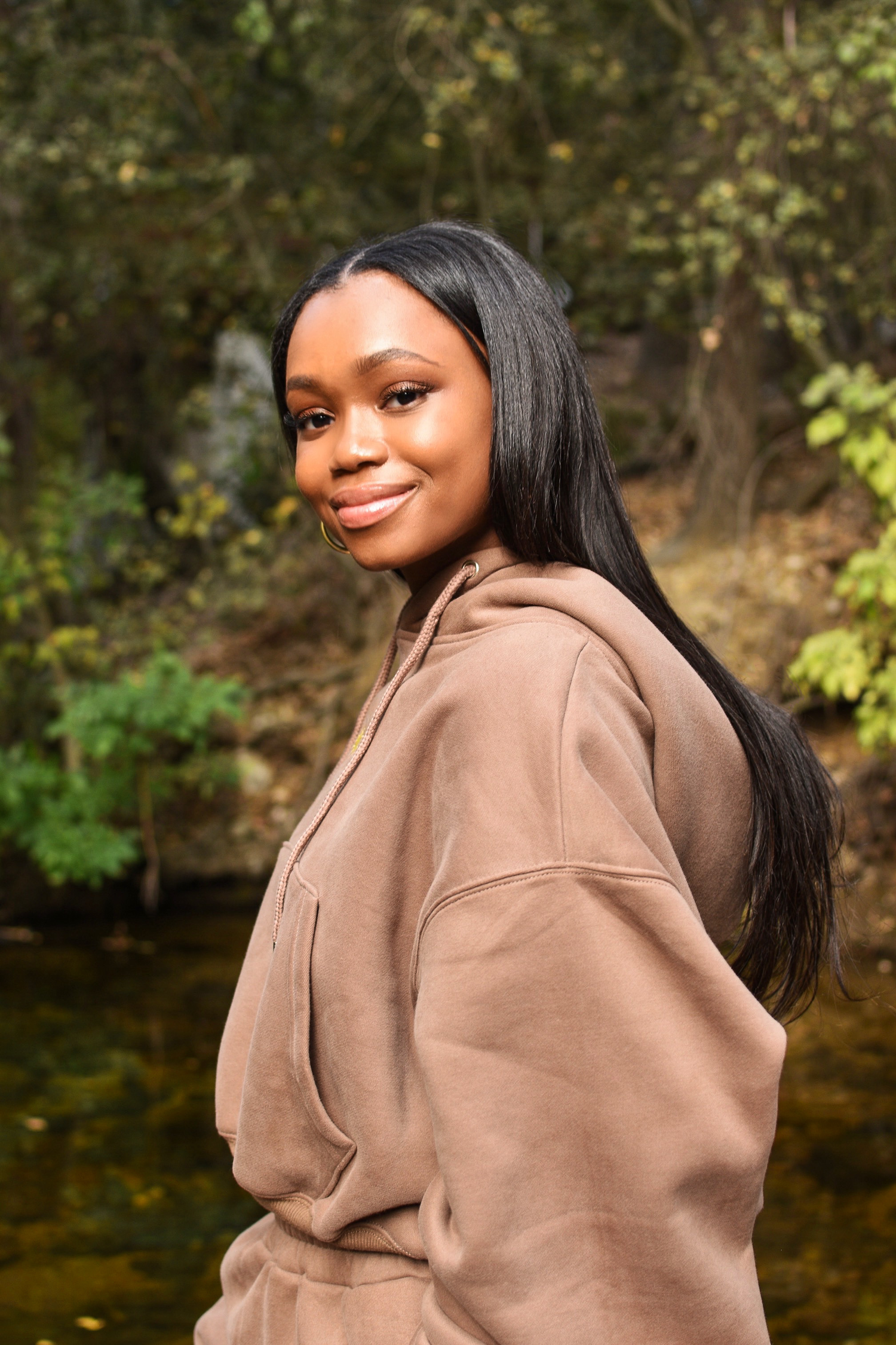 A person in a brown hoodie smiles while posing outdoors near a pond.