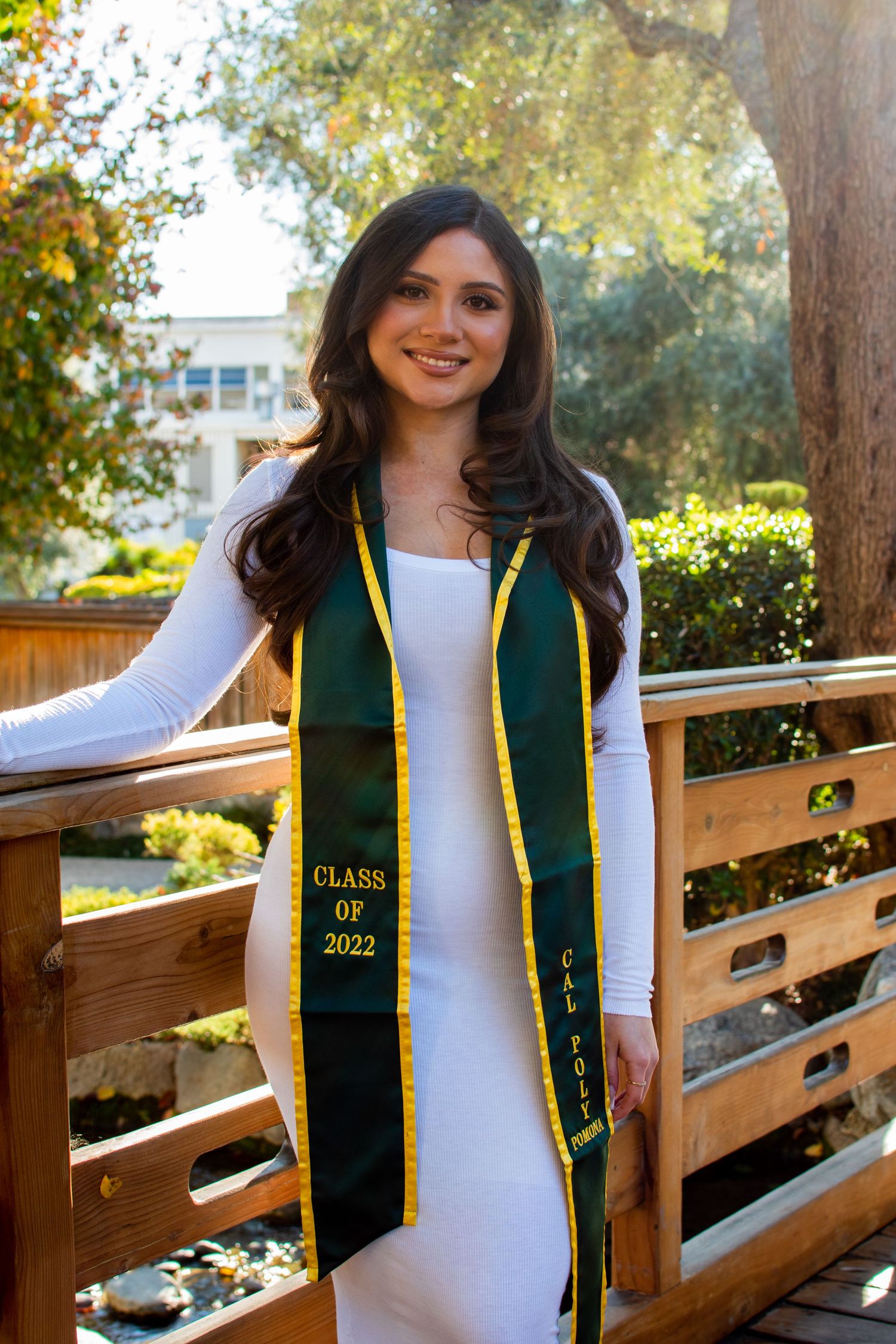 Graduate in white dress wearing green and gold graduation stole posing on wooden bridge outdoors in sunny garden setting.