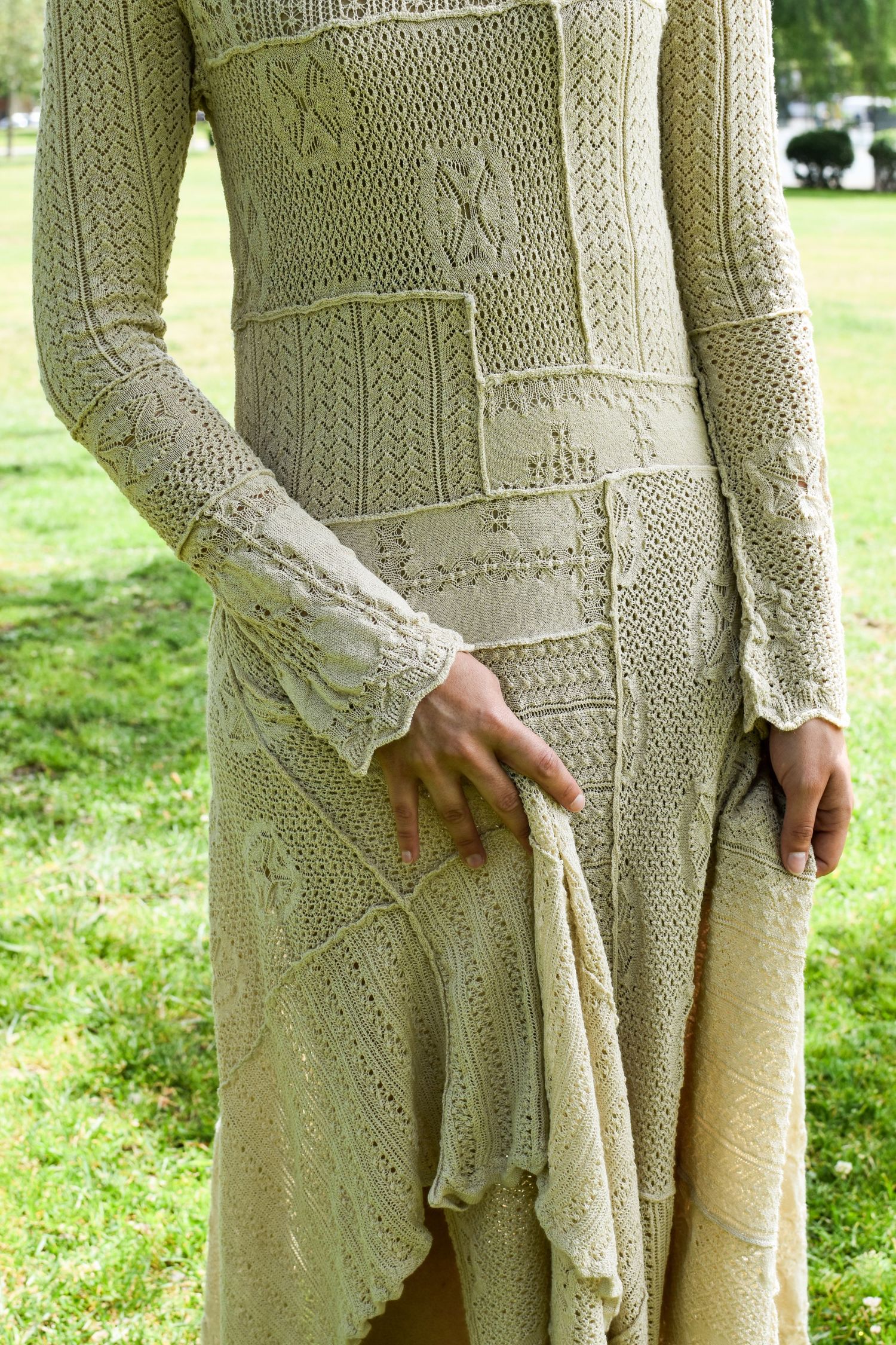 Close-up detail of an intricate cream-colored knit dress with cable and lace patterns on a grassy background.