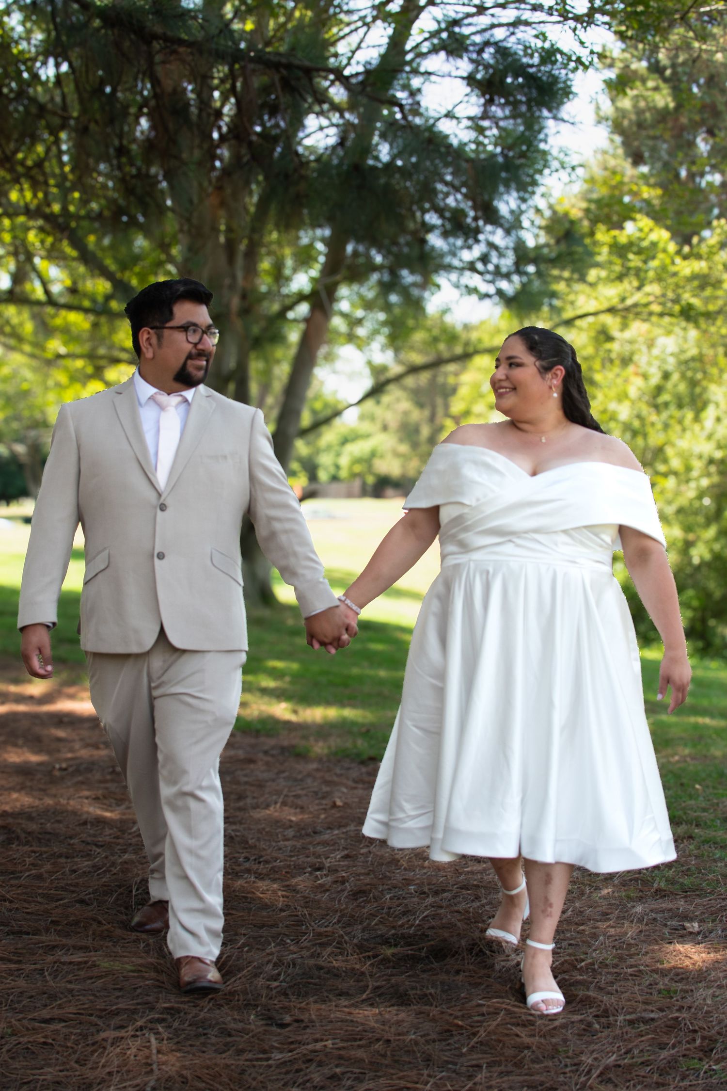 A couple in wedding attire walk hand in hand along a tree-lined path on their special day.