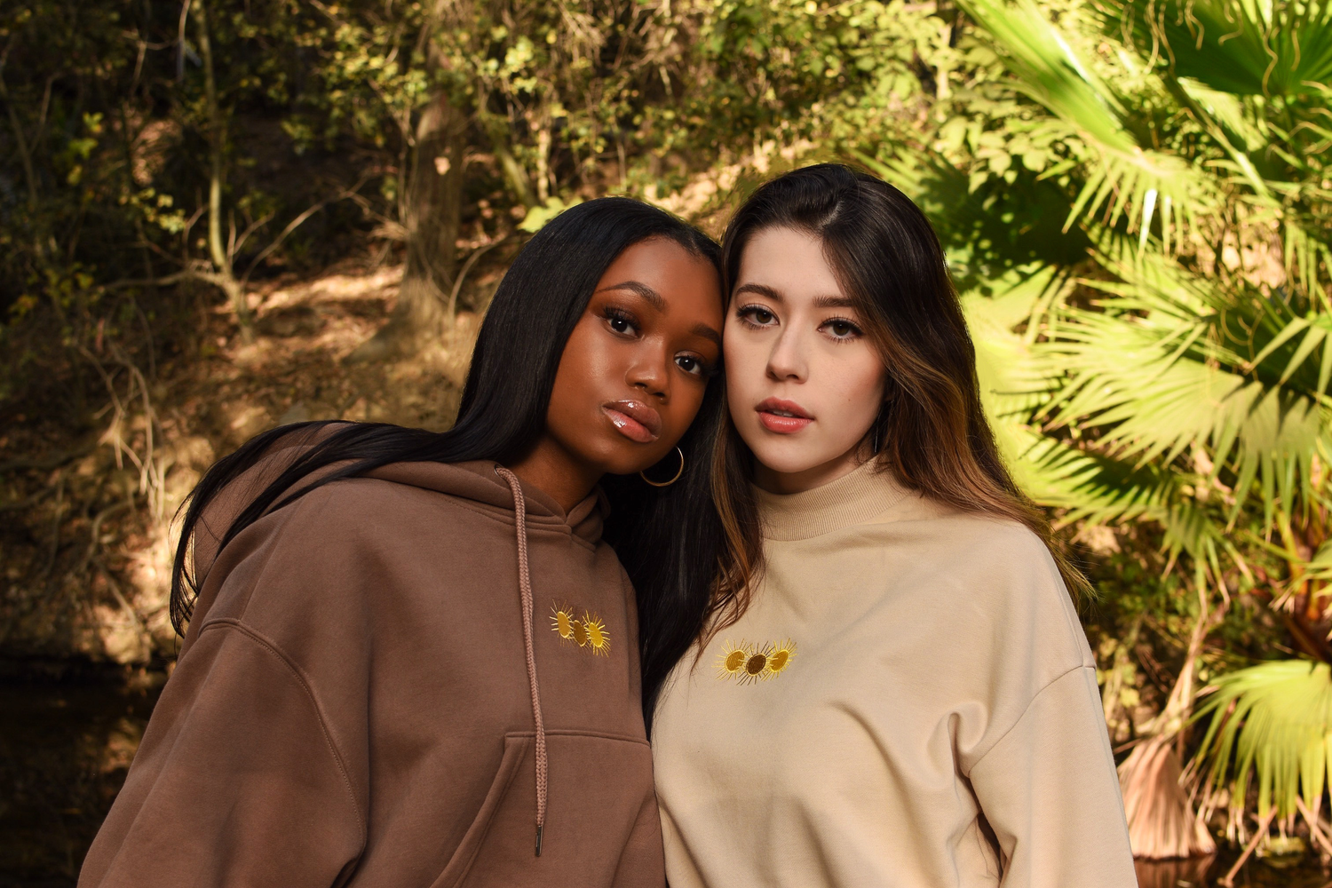 Two models in matching hooded sweatshirts pose together against a tropical foliage background.