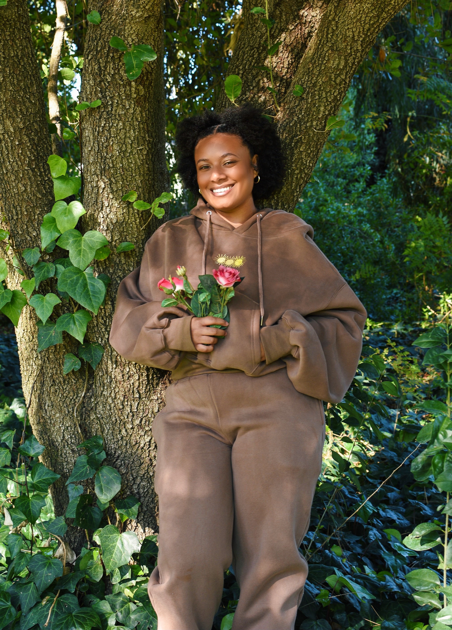 Woman in brown loungewear holding flowers while standing against tree trunk outdoors.