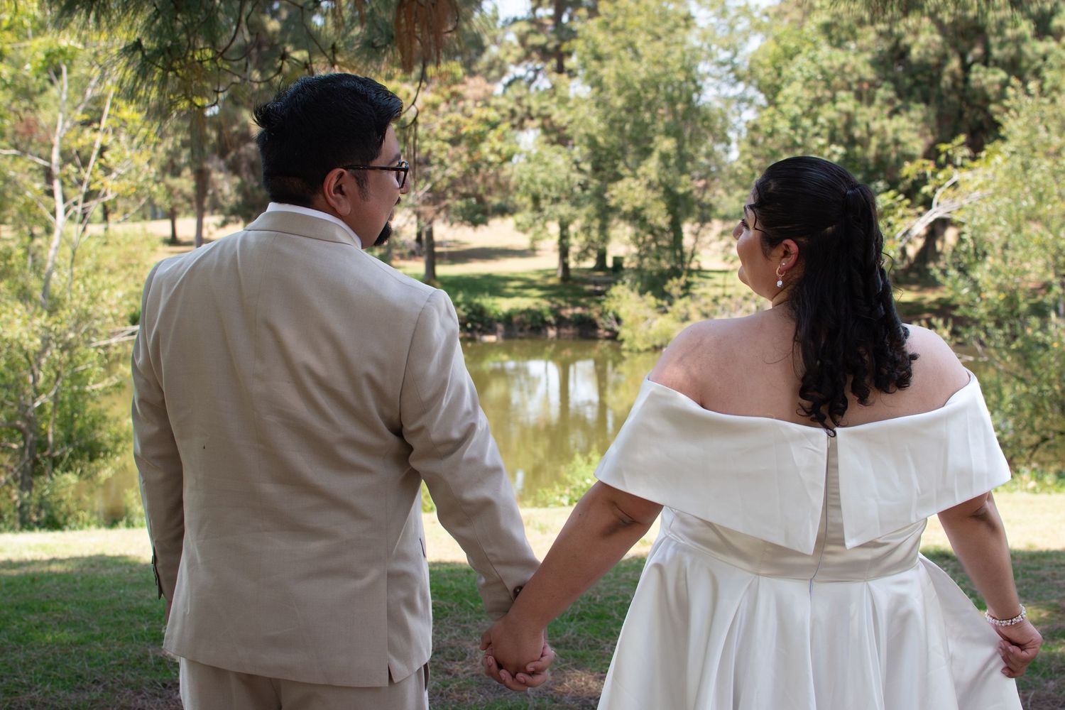 A couple in white and beige wedding attire holds hands while looking out over a serene pond in a garden setting.