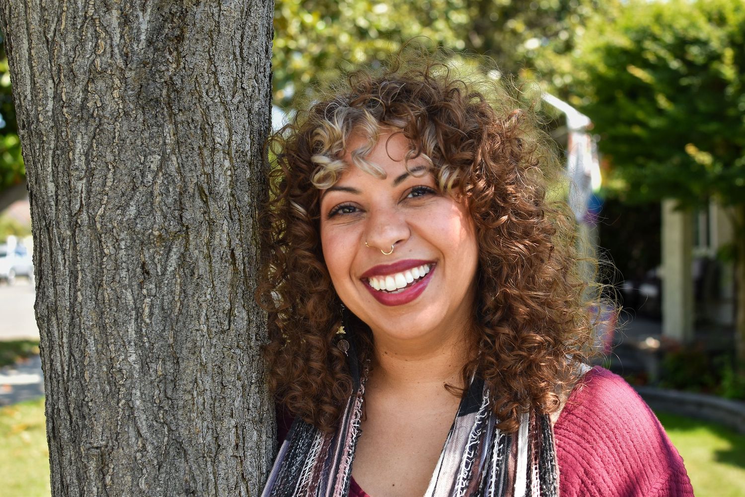 Person with curly brown hair and bright smile leaning against tree trunk in outdoor setting on sunny day.