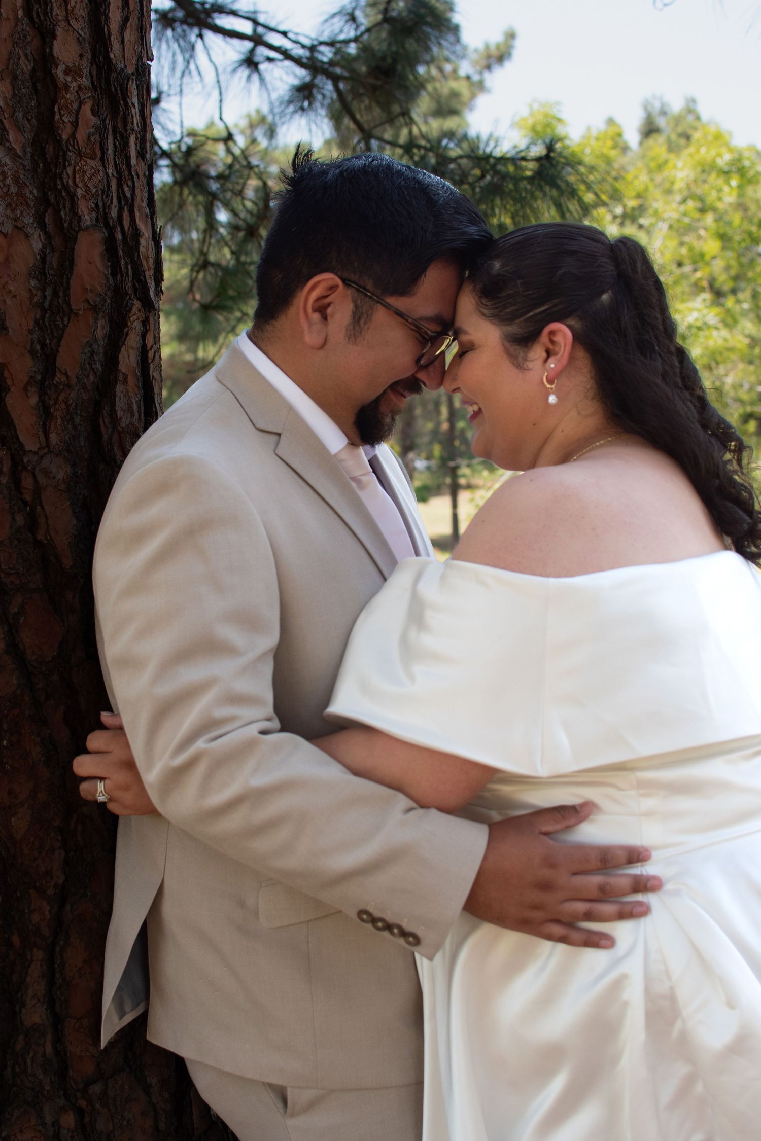 Happy newlyweds embrace and laugh together in a garden setting wearing coordinated beige and white wedding attire.