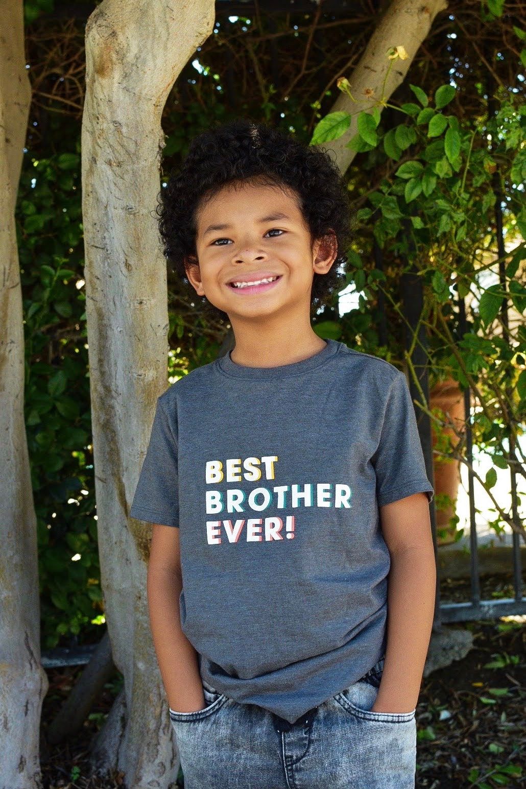 Child wearing a gray t-shirt with 'Best Brother Ever' text posing against a tree outdoors.