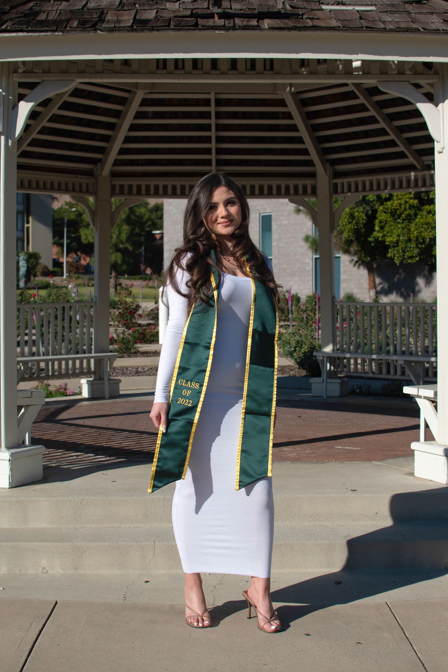 Graduate in commencement regalia sits in white gazebo during sunny photoshoot.