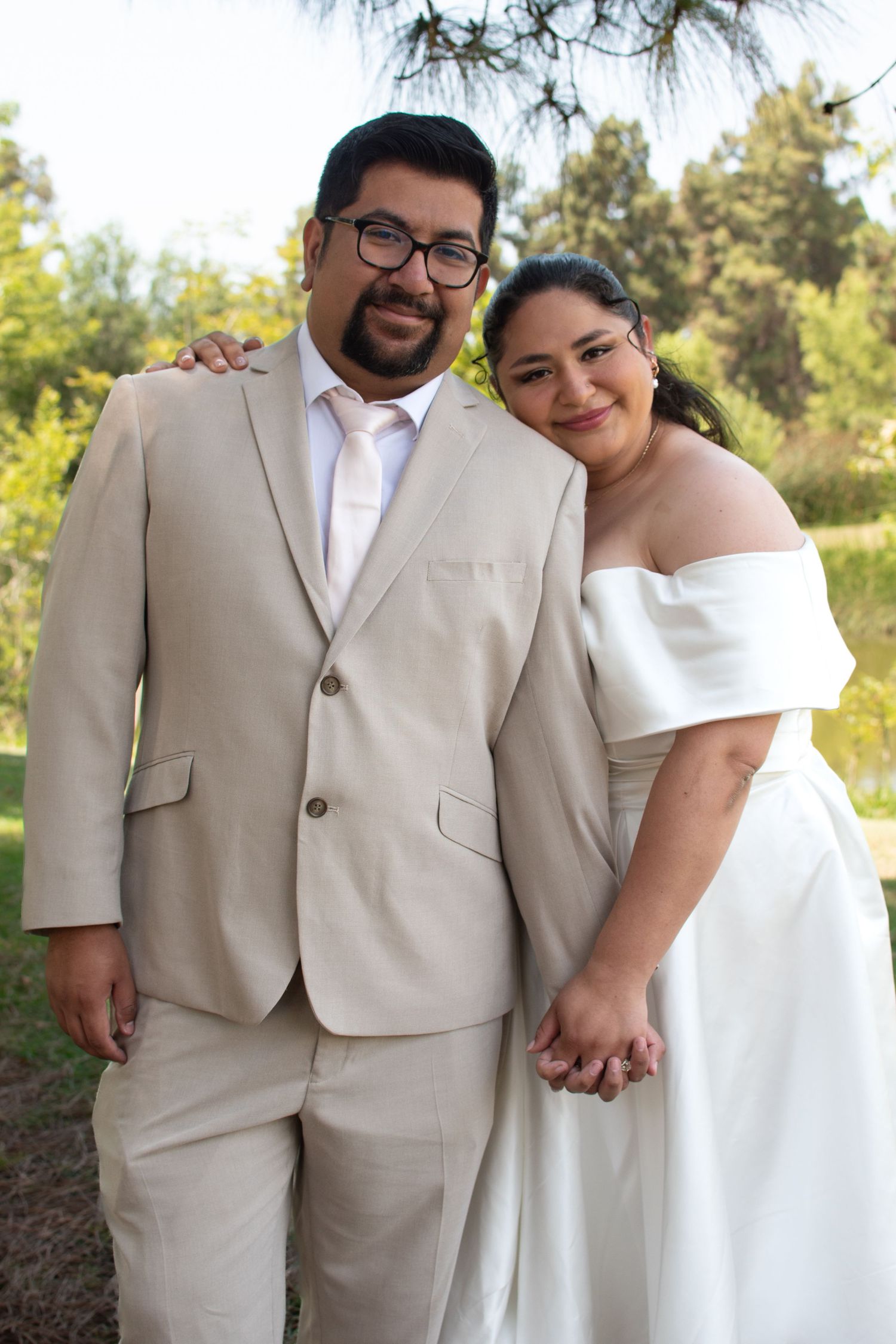 Couple in outdoor wedding attire with tan suit and white off-shoulder dress pose together against natural backdrop.