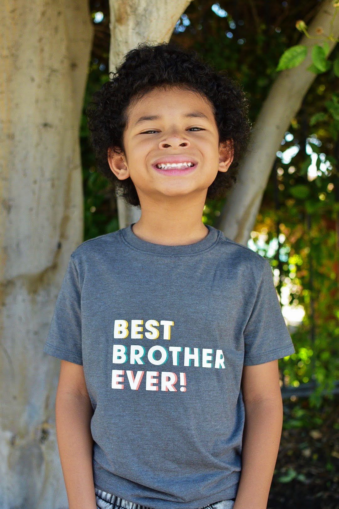 Child in 'Best Brother Ever' t-shirt smiles cheerfully while standing against a tree.
