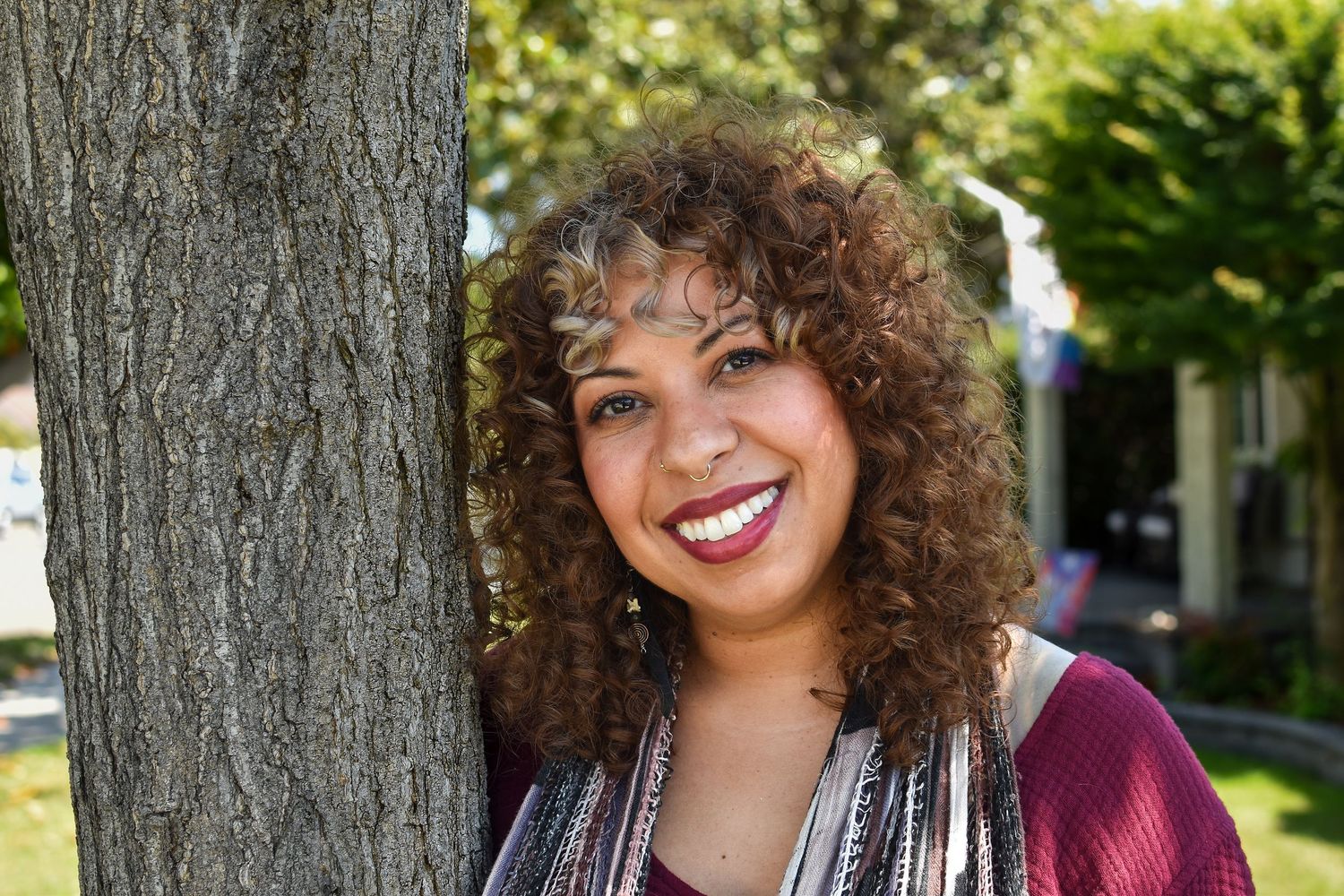 Casual outdoor portrait of a person with curly hair wearing a burgundy top leaning against a tree on a sunny day.