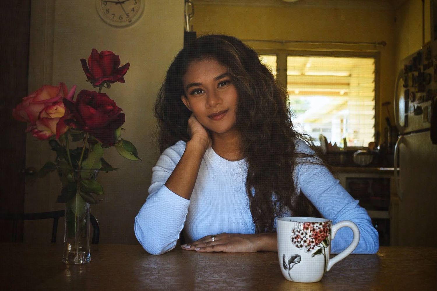 Person sitting at wooden table with decorative mug and red roses in vase.