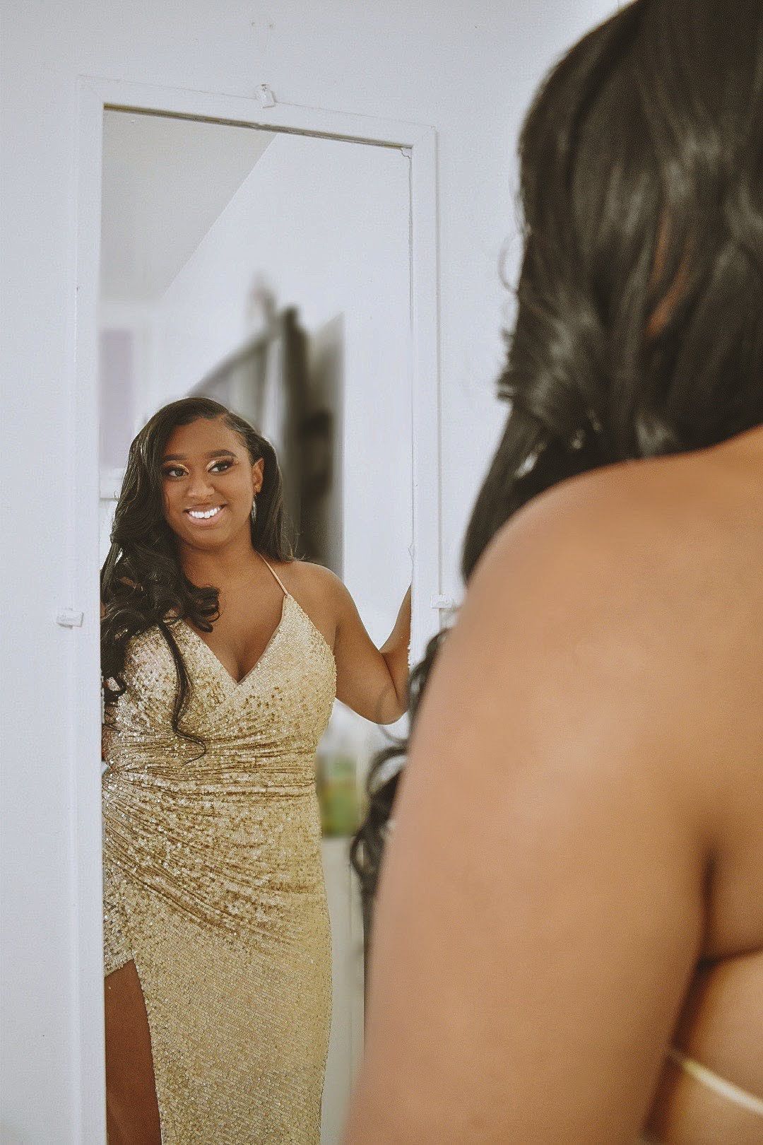 Person in gold sequin gown smiling at reflection in full-length mirror.