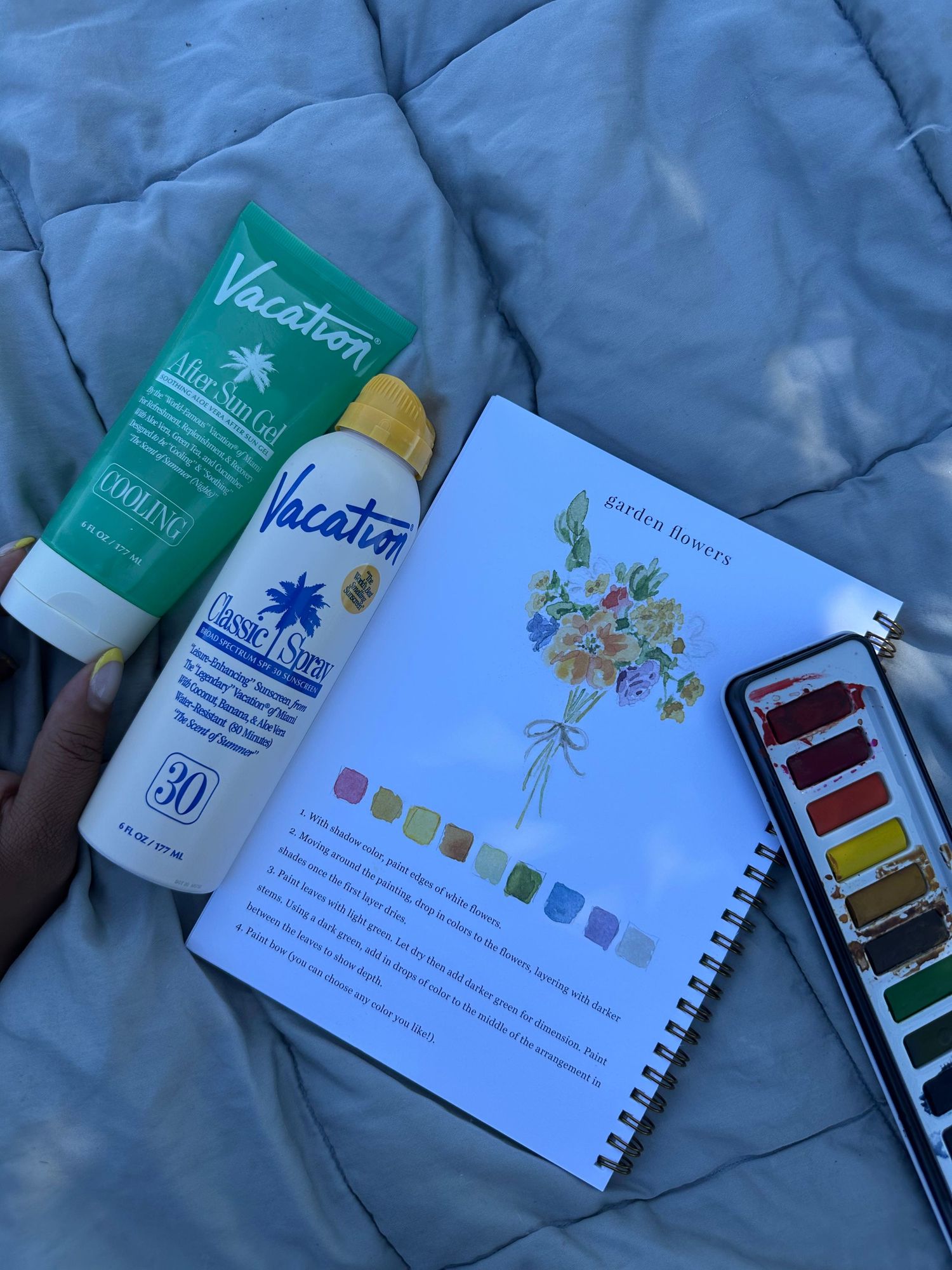 Art supplies including sunscreen, sketchbook with floral illustration, and watercolor palette on blue fabric.