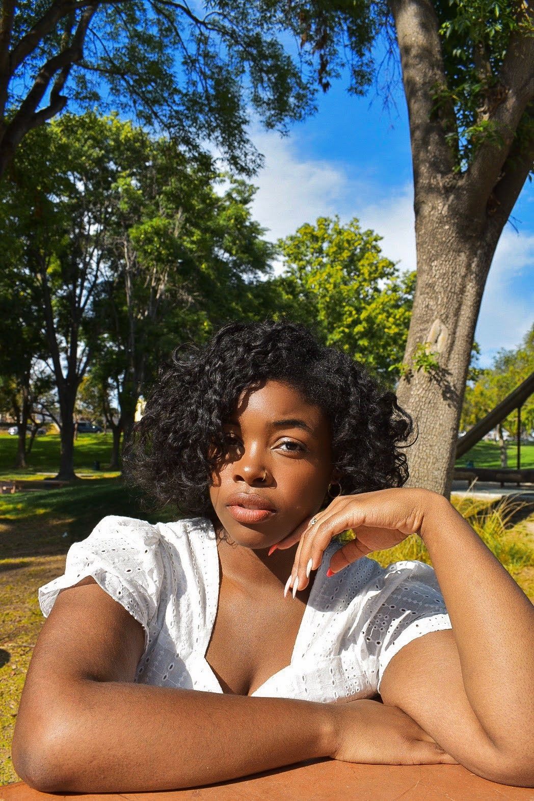 Outdoor portrait at park with white eyelet top against green tree background.