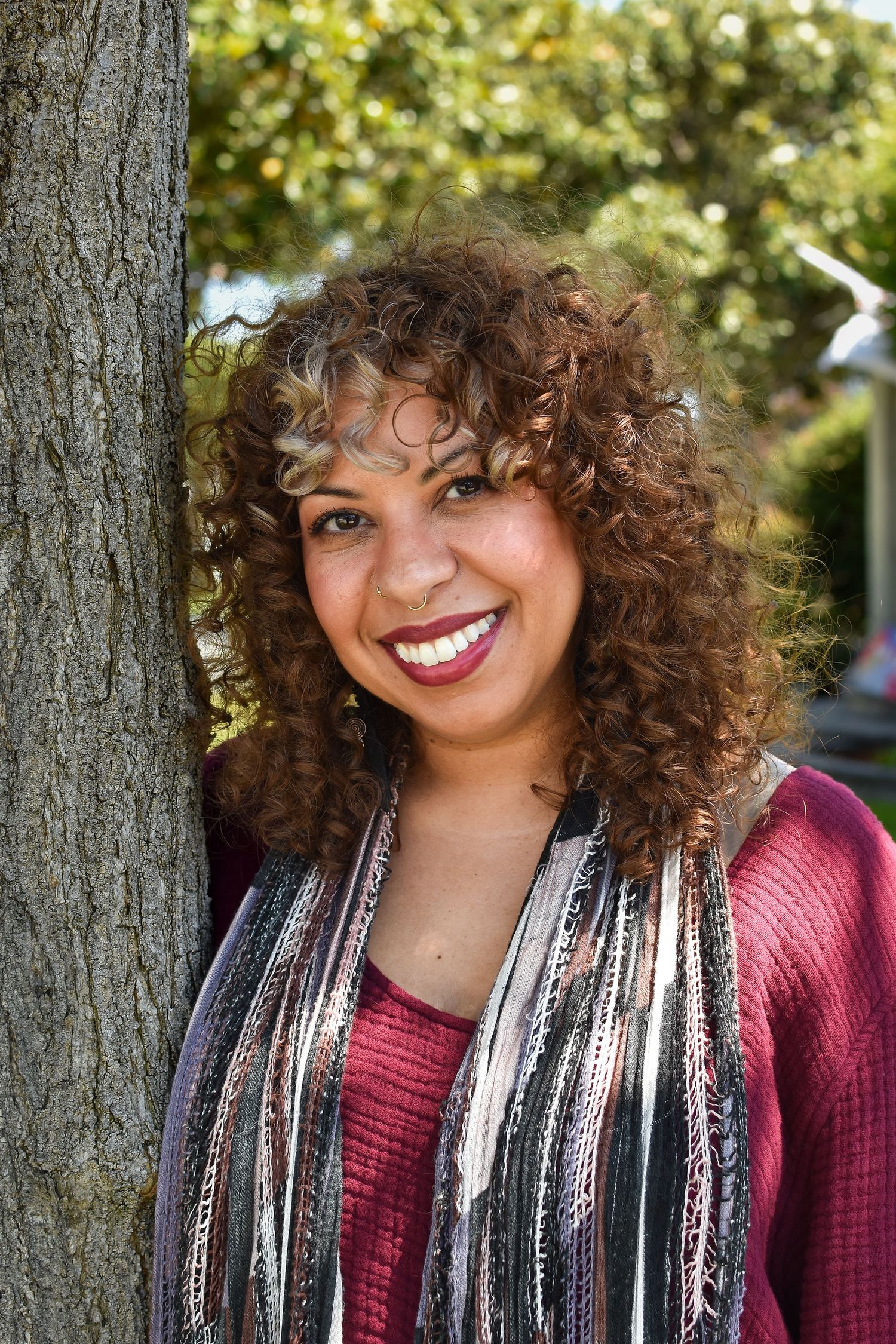Curly haired person wearing a burgundy sweater and striped scarf smiles warmly against a sun-dappled leafy background.