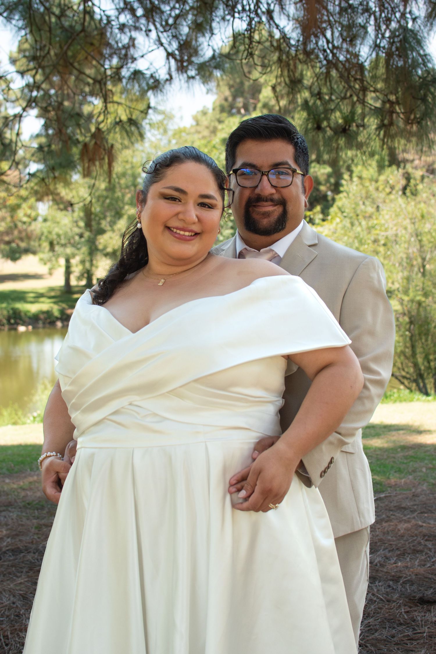 A couple poses outdoors on their wedding day wearing a white gown and beige suit near a pond with trees in the background.