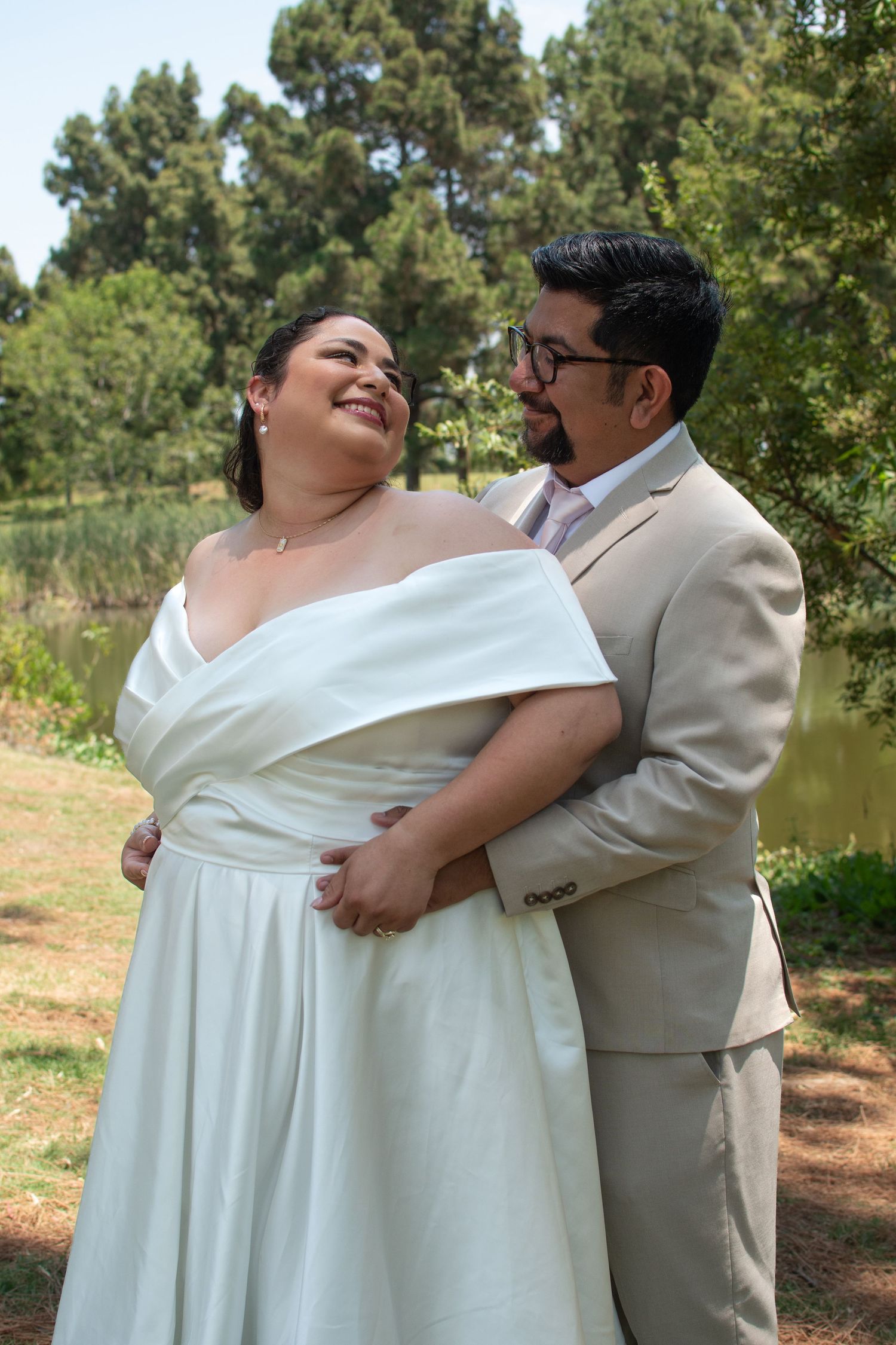 Couple shares a joyful moment outdoors in wedding attire, with a white dress and beige suit against a natural backdrop.