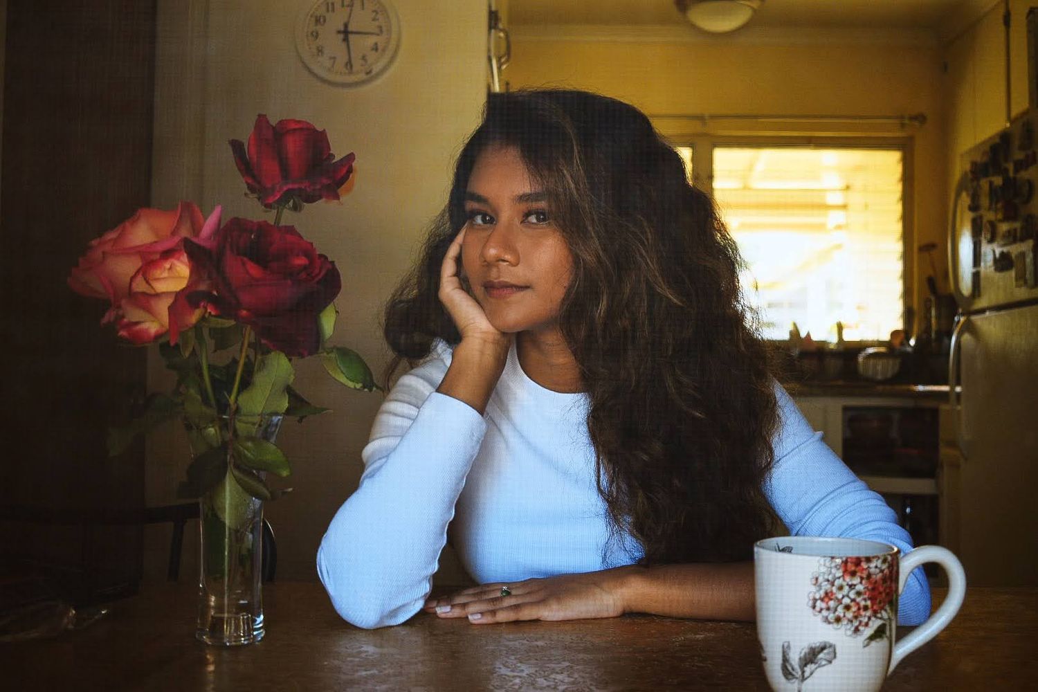 Person resting at wooden table with floral mug and red roses in soft lighting.