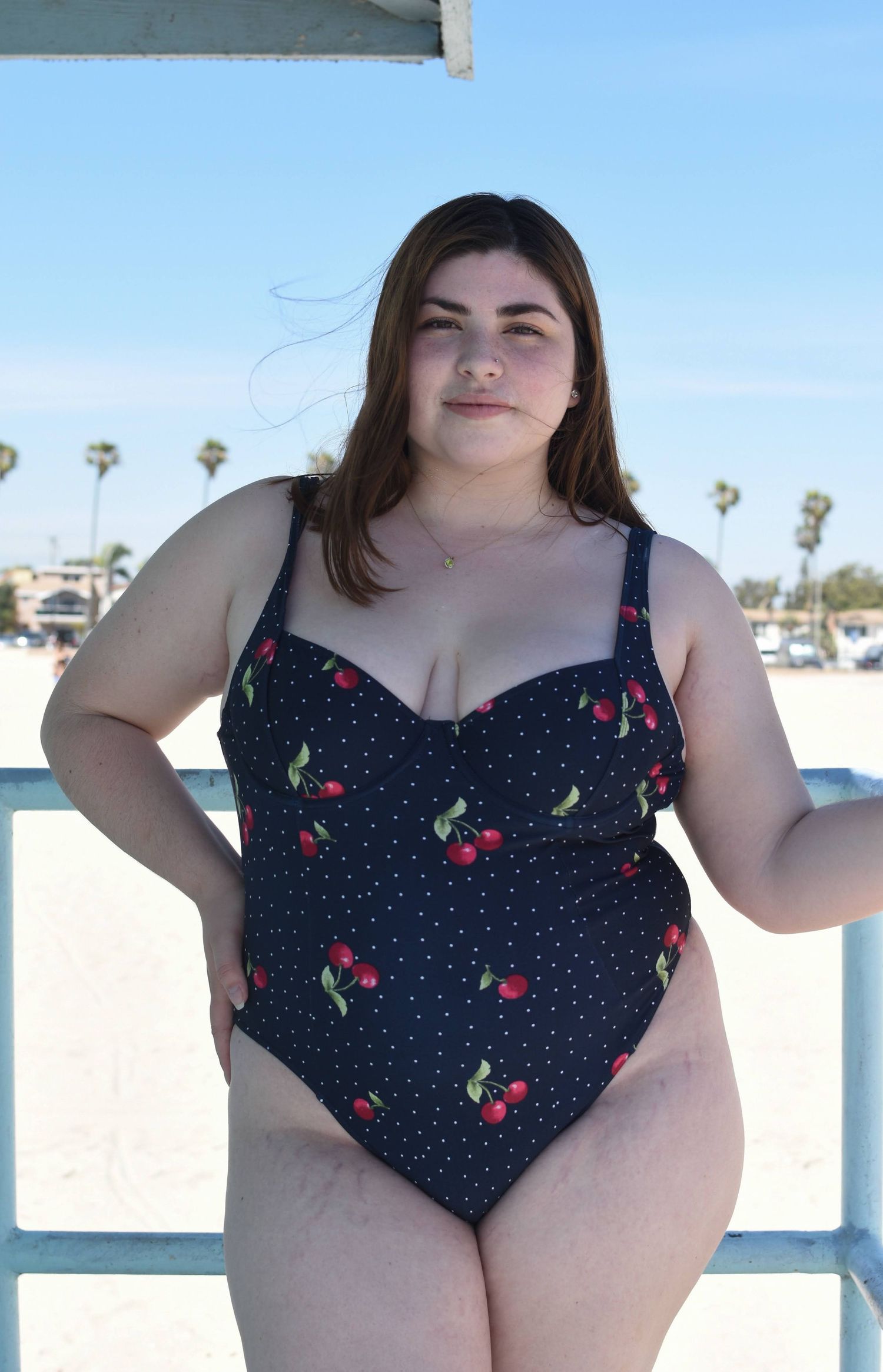 A navy one-piece swimsuit with cherry and polka dot pattern shown against a beach backdrop.