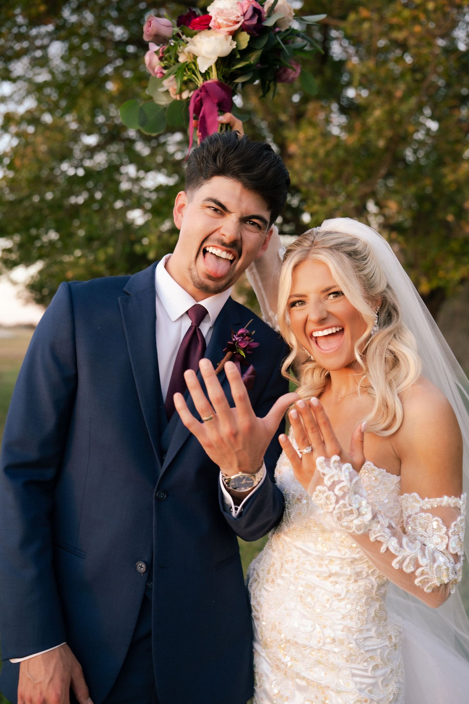 Joyful newlyweds showing off wedding rings, laughing outdoors with colorful floral arch behind them.