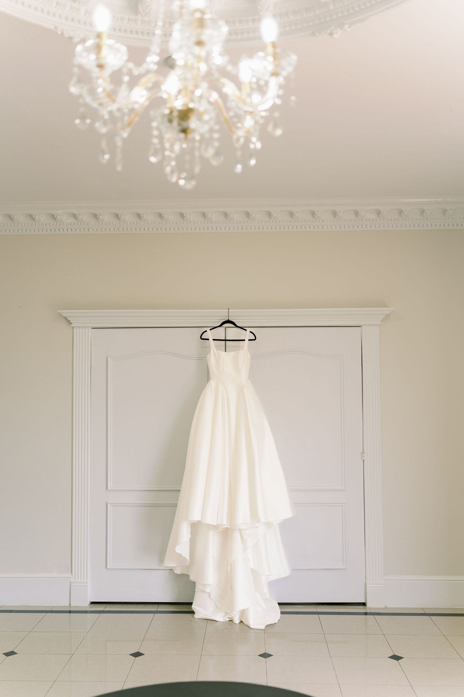 Elegant strapless white wedding gown hanging on hanger beneath crystal chandelier in white room.