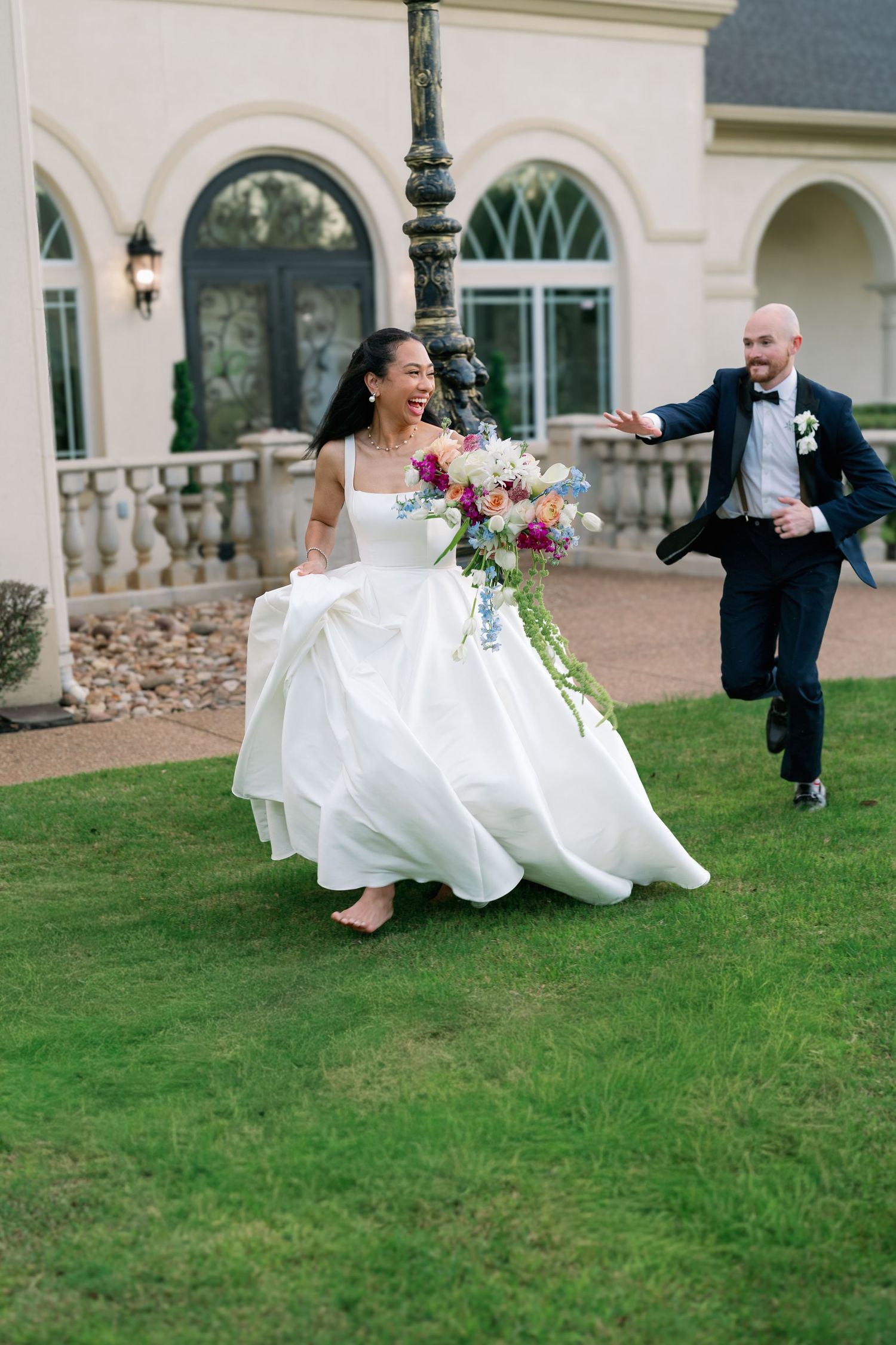 Joyful bride in white gown running with bouquet as groom chases her outdoors.