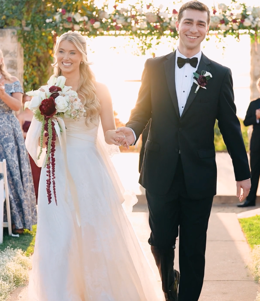 Newlyweds walk down aisle hand in hand, bride holding white and burgundy bouquet.
