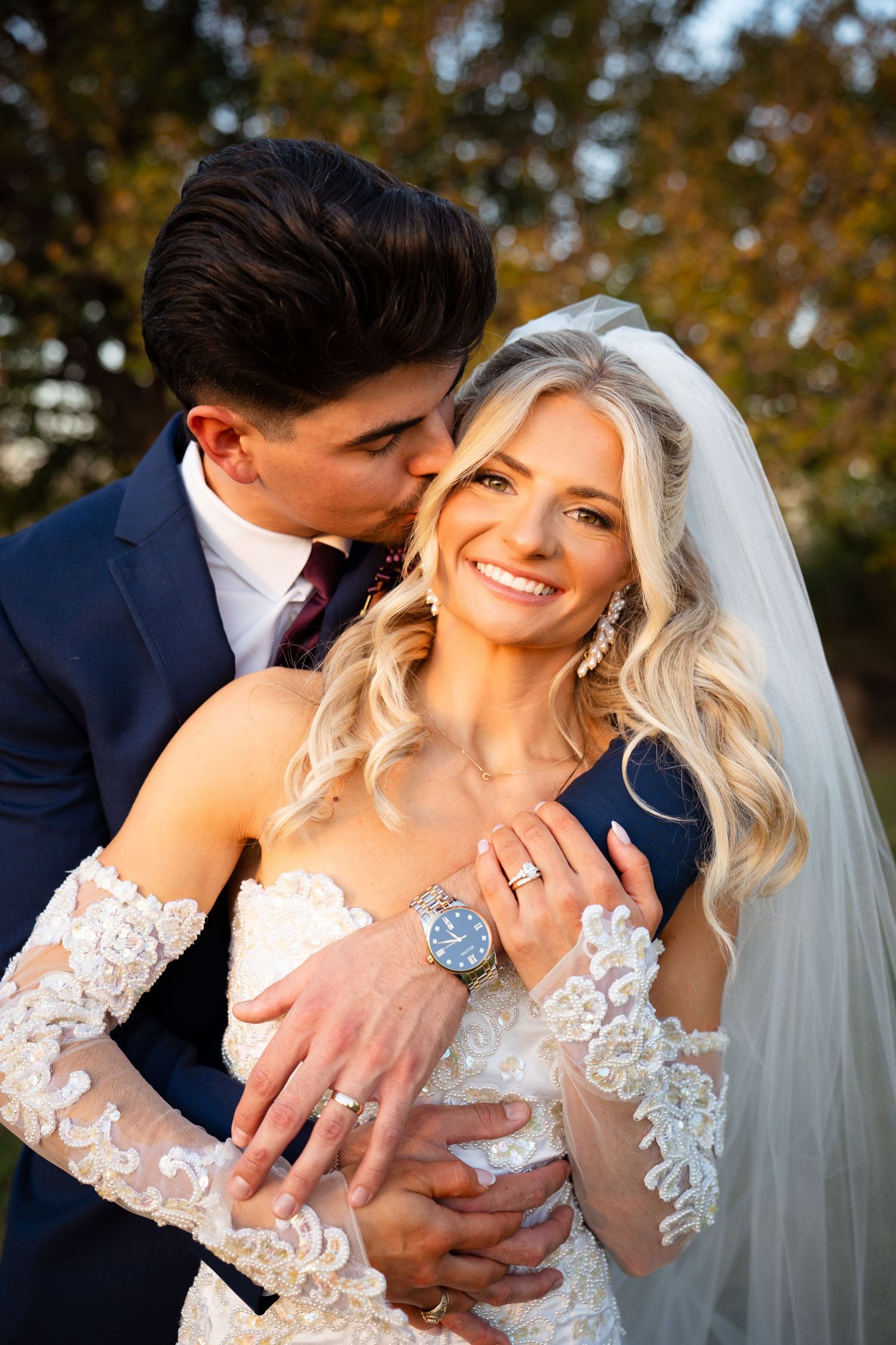 Groom kissing bride on cheek while she smiles, both wearing wedding rings in autumn setting.